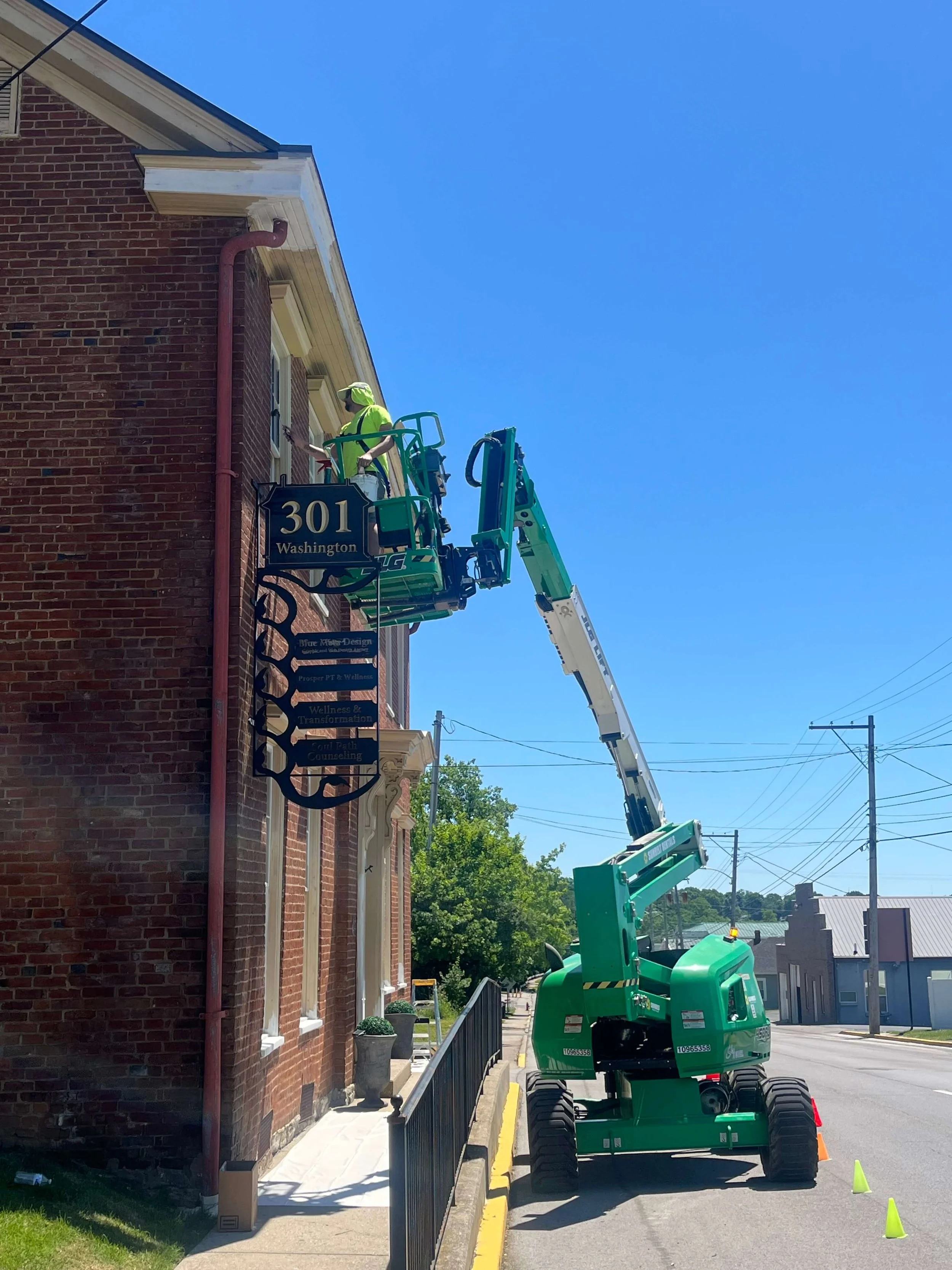 A worker on a green cherry picker working on the exterior of a brick building at 301 Washington. The worker is wearing a yellow safety helmet and high-visibility vest, and is using a tool. There are small orange cones set up around the cherry picker on a city street with power lines overhead and other buildings visible in the background.