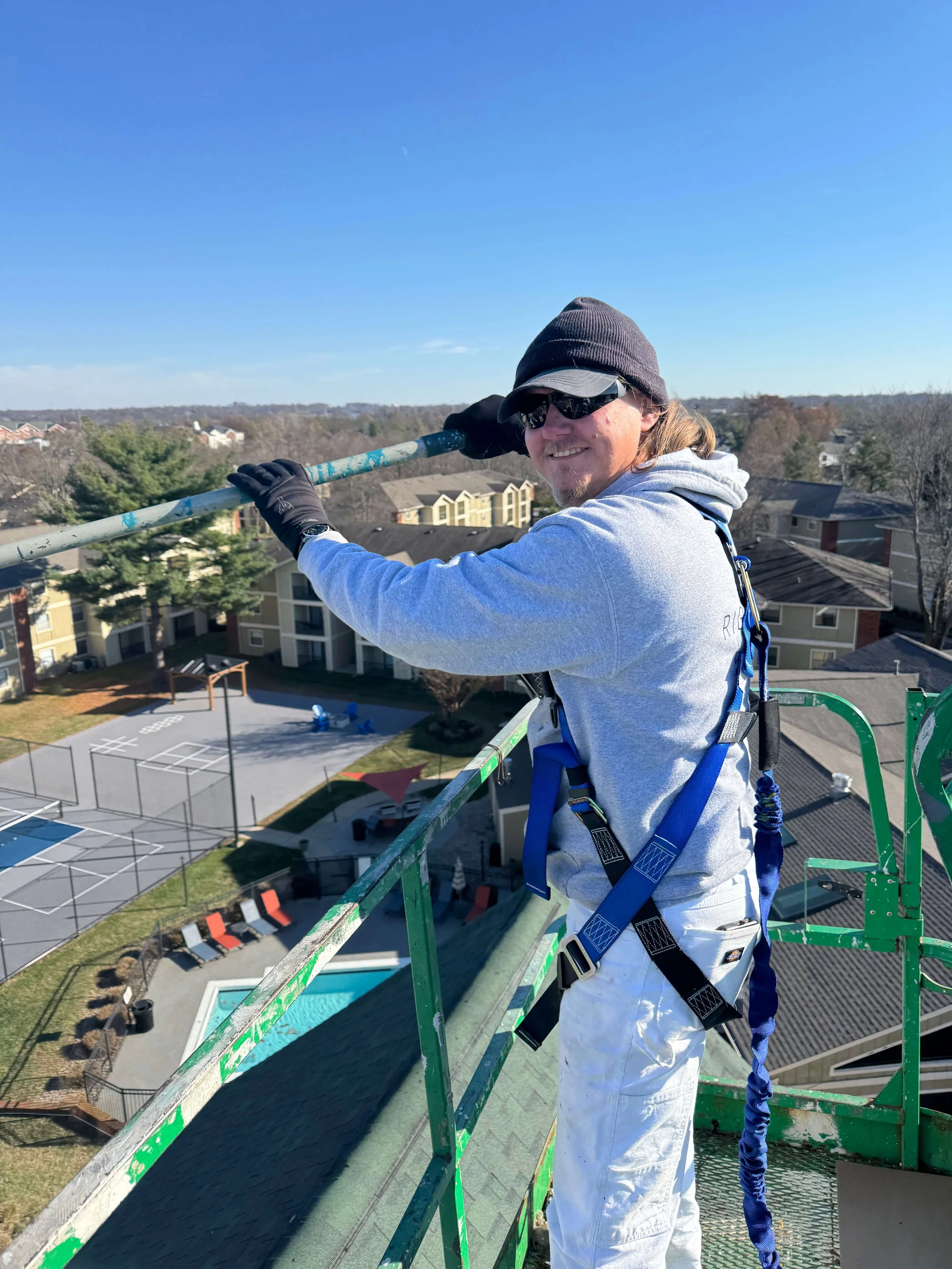 A man wearing a gray hoodie, black gloves, sunglasses, and a gray beanie hat standing on a lift platform, smiling and holding onto a pole, with a background of trees, apartment buildings, a basketball court, and a swimming pool under a clear blue sky.