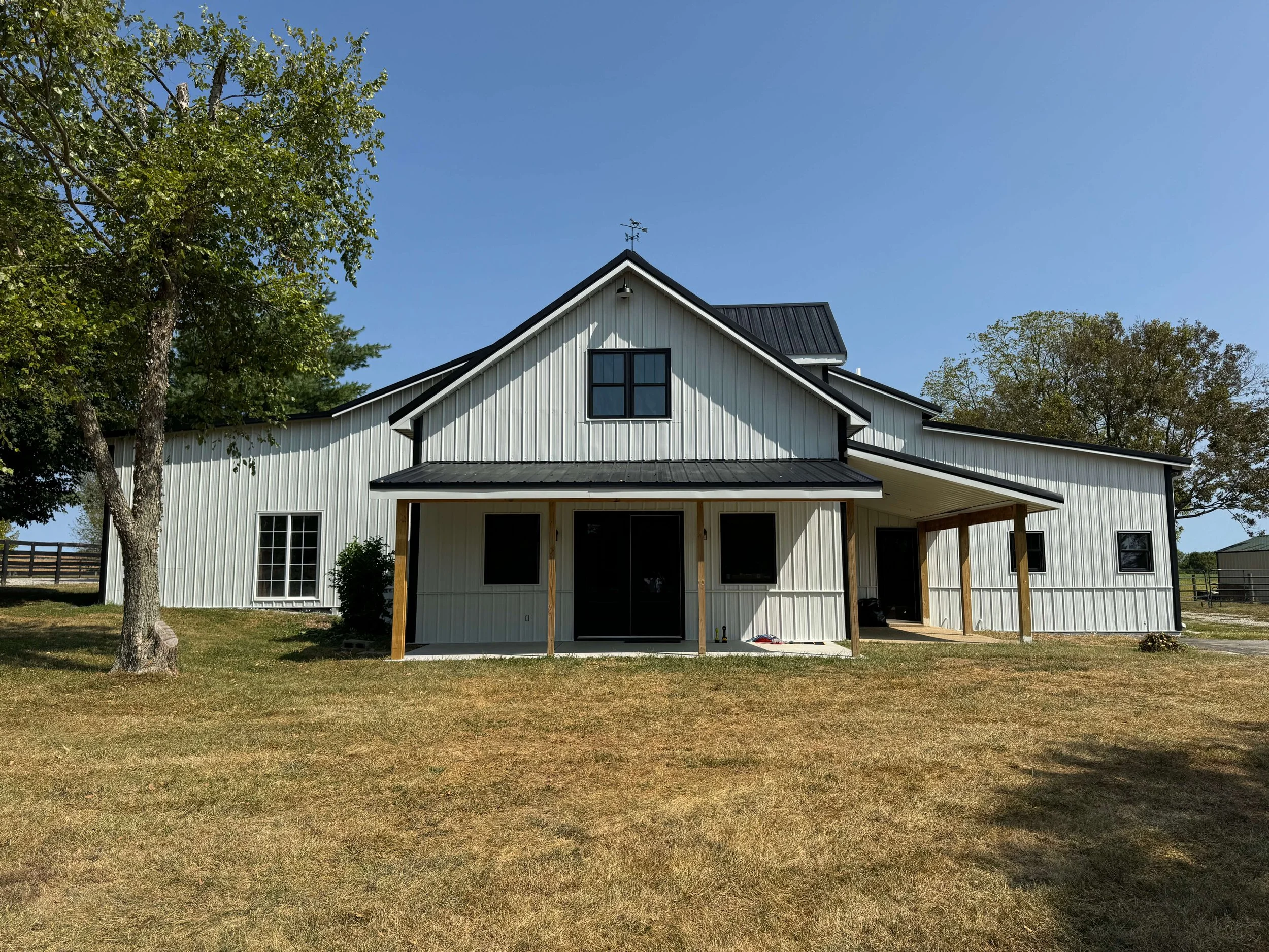 White barn with black roof and a weathervane, surrounded by trees and green grass, under a clear blue sky.