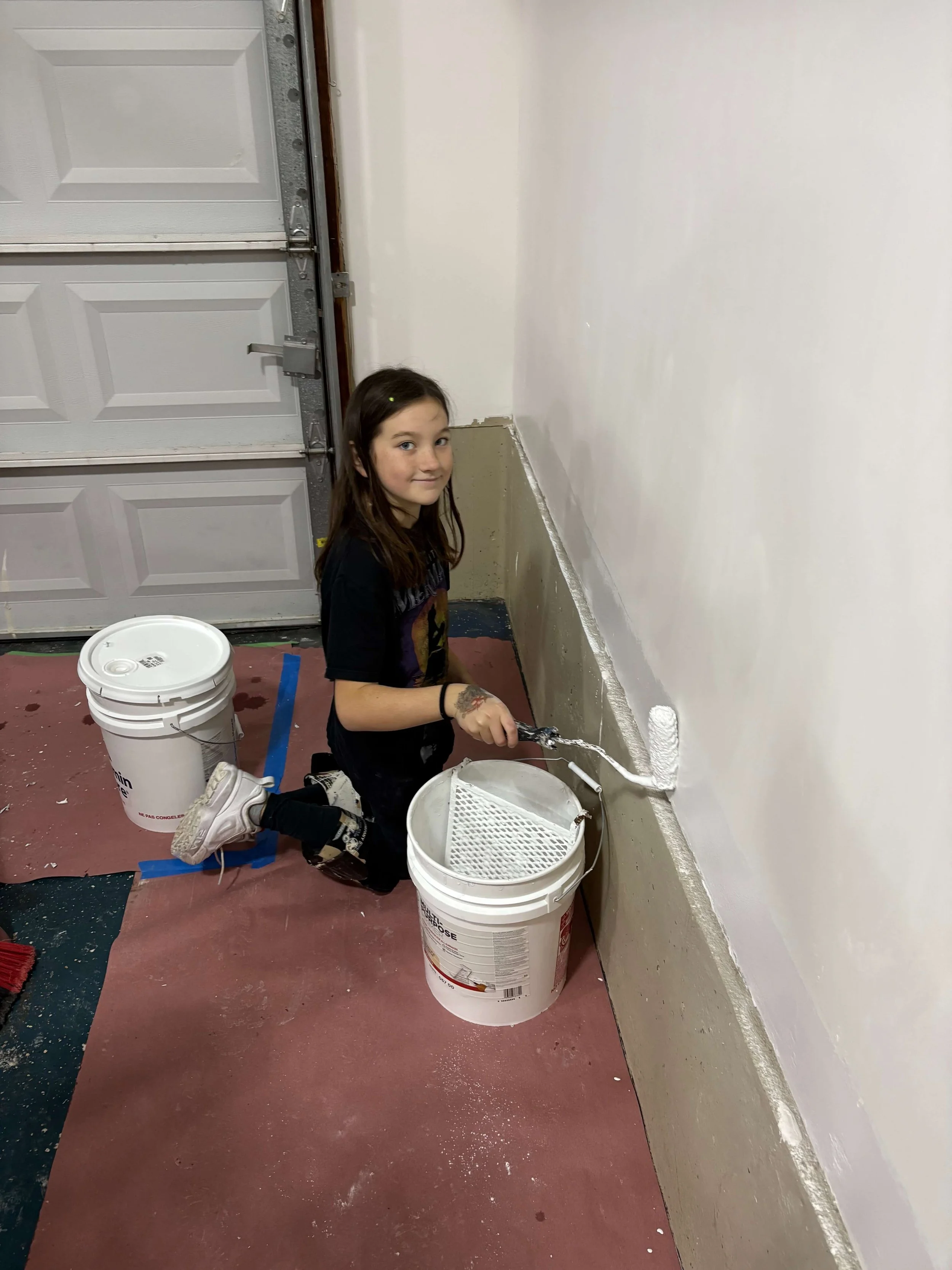 A girl kneeling on the floor painting a baseboard white in a garage or utility room, with buckets of white paint around her.