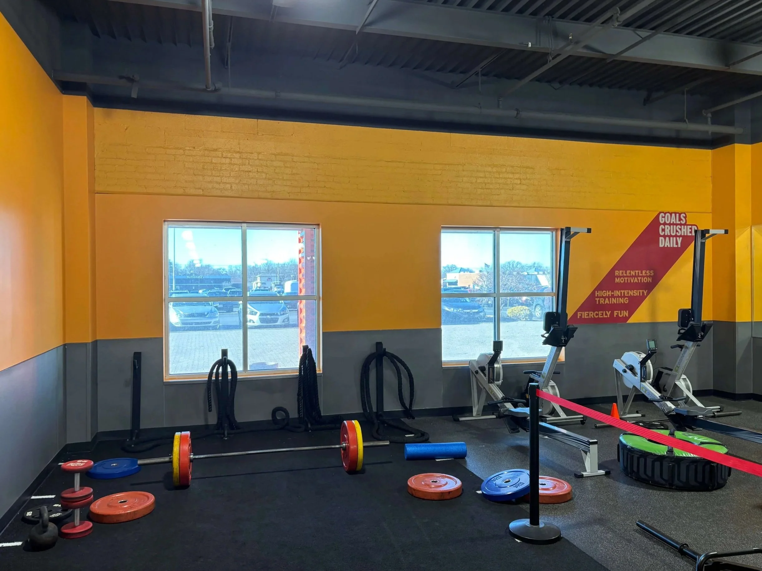Interior of a gym with weightlifting equipment, barbells, weight plates, and exercise machines, with yellow walls, large windows, and gym motivational poster.