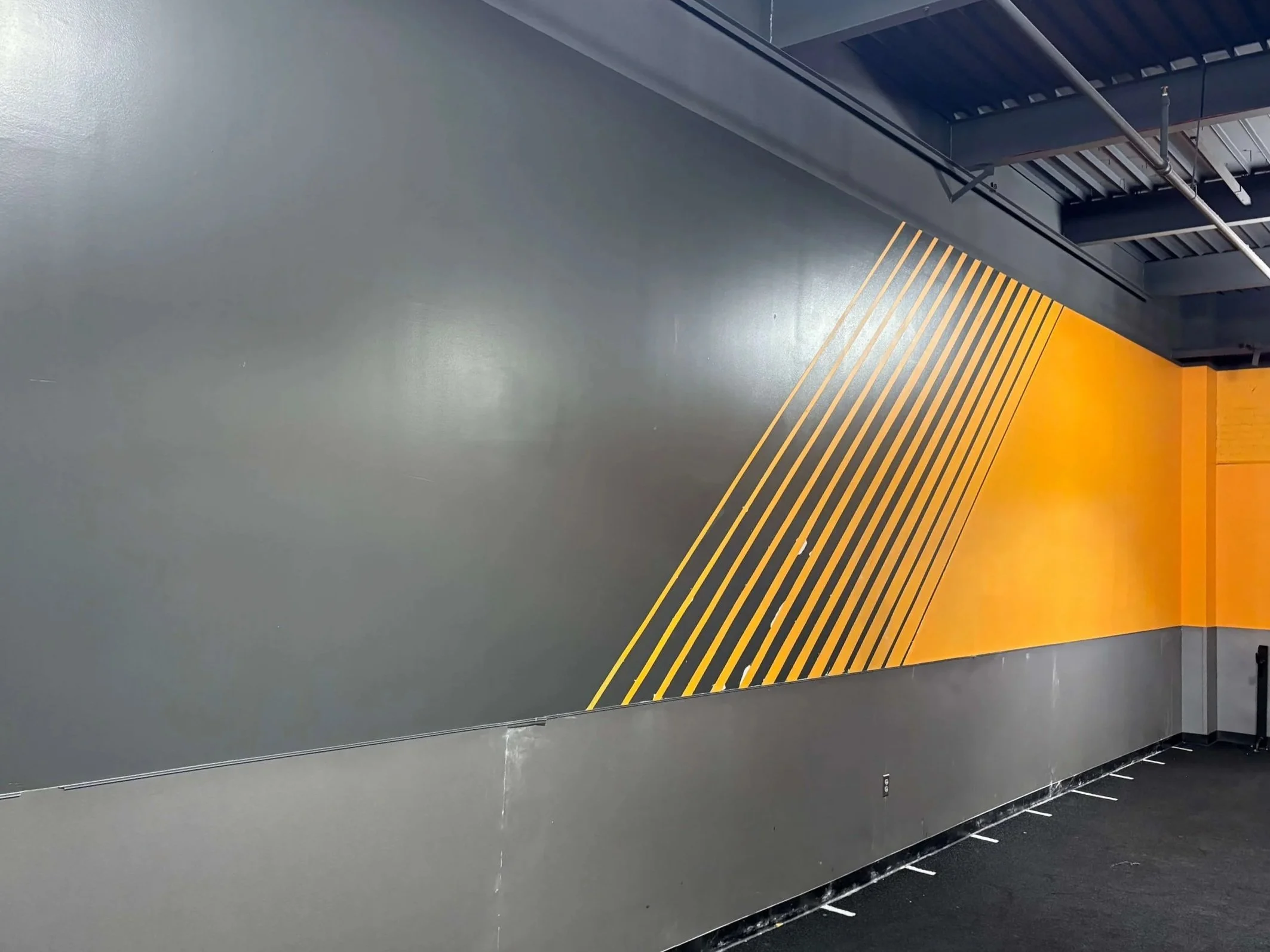 A modern interior wall with a gray section and a yellow diagonal striped section, with metal pipes and a black ceiling above.