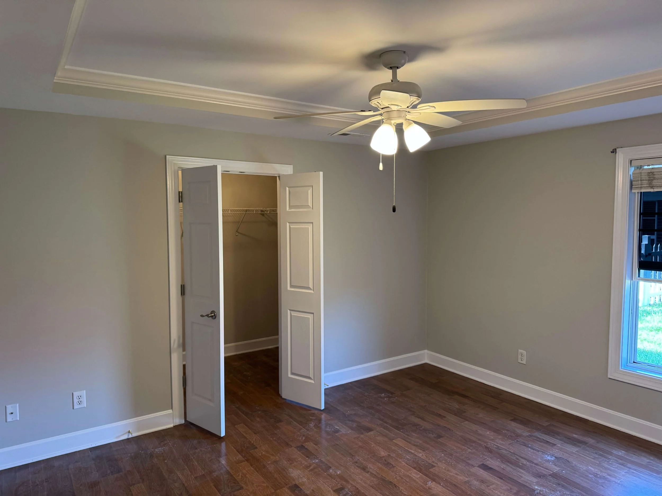 Empty room with hardwood flooring, light-colored walls, a ceiling fan with lights, a window with blinds, and an open closet with bifold doors.