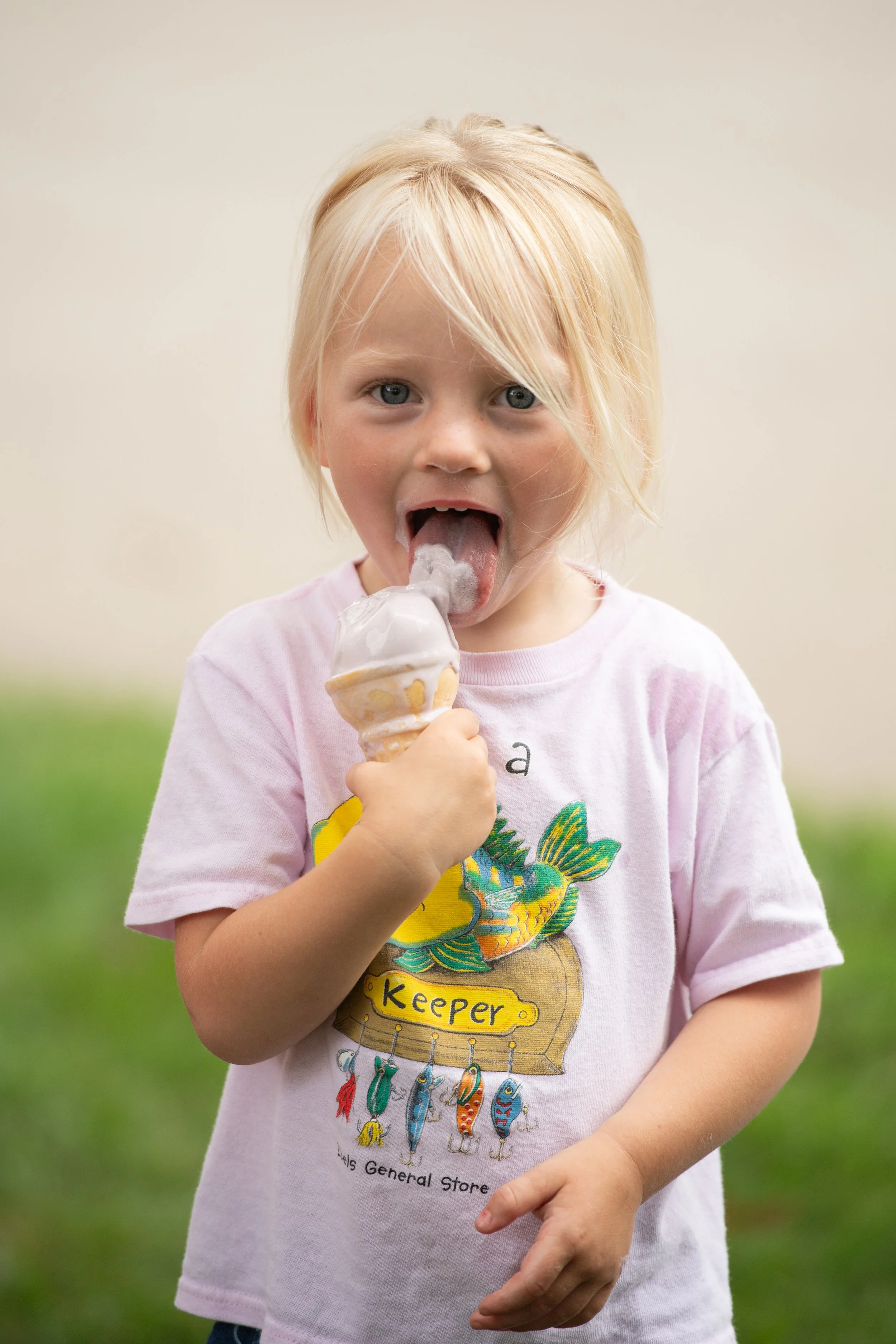 A young girl with blonde hair and blue eyes is licking a vanilla ice cream cone outside in a grassy area, wearing a pink T-shirt with a colorful fish and fishhook design.