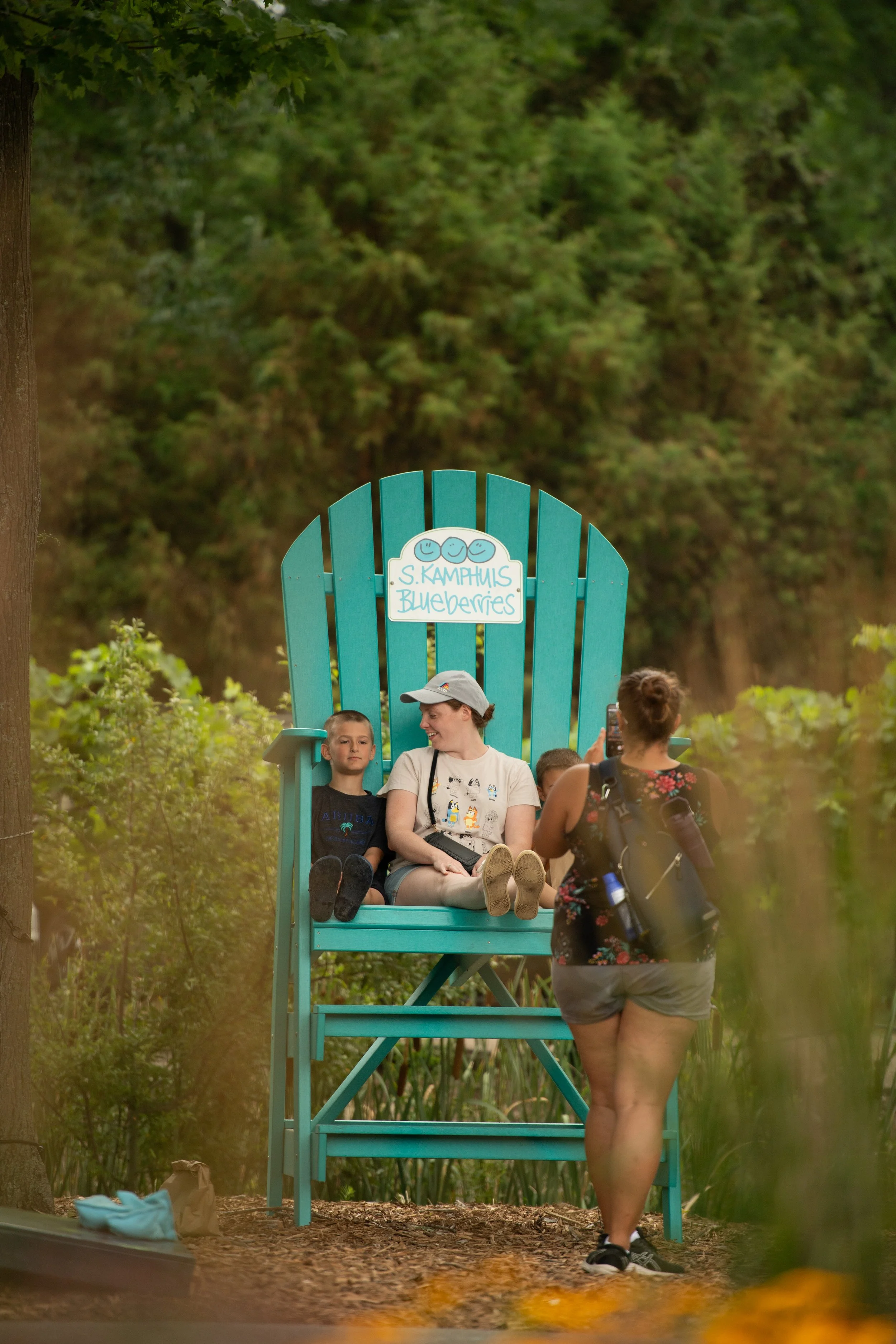 A woman taking a photo of a boy and a woman sitting on an oversized beach chair labeled 'S. Kampmans Blueberries' in a green outdoor setting.