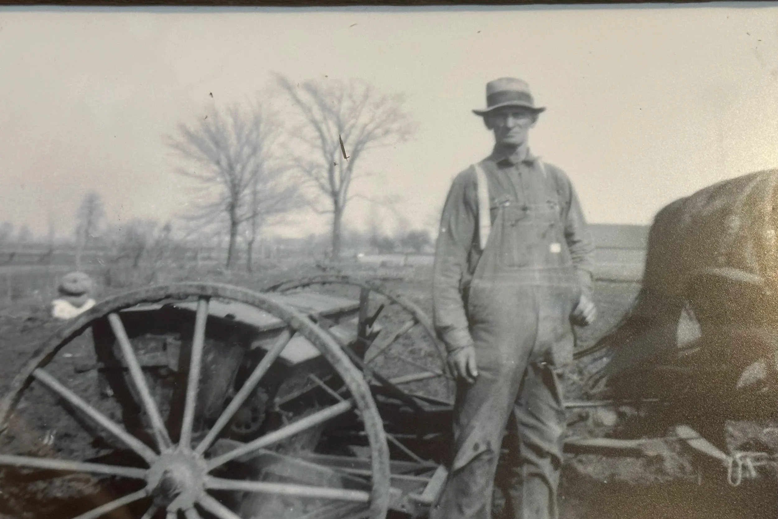 A black and white photo of a man in work clothes and a hat standing outdoors with a large wooden wheel wagon, trees, and a second person in the background.