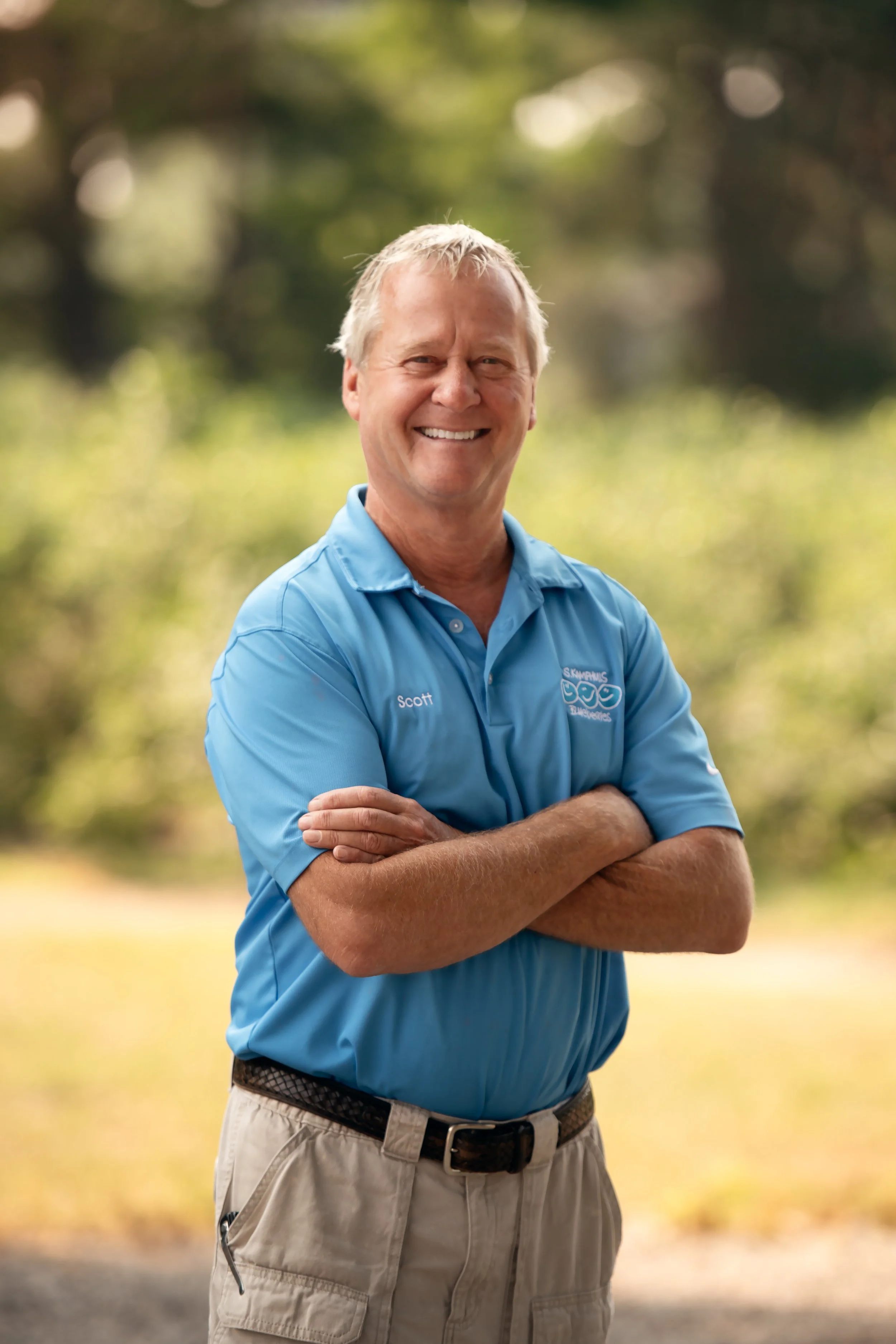 A smiling middle-aged man with gray hair standing outdoors with his arms crossed, wearing a blue polo shirt with the name 'Scott' and a logo, in front of a blurred green background.
