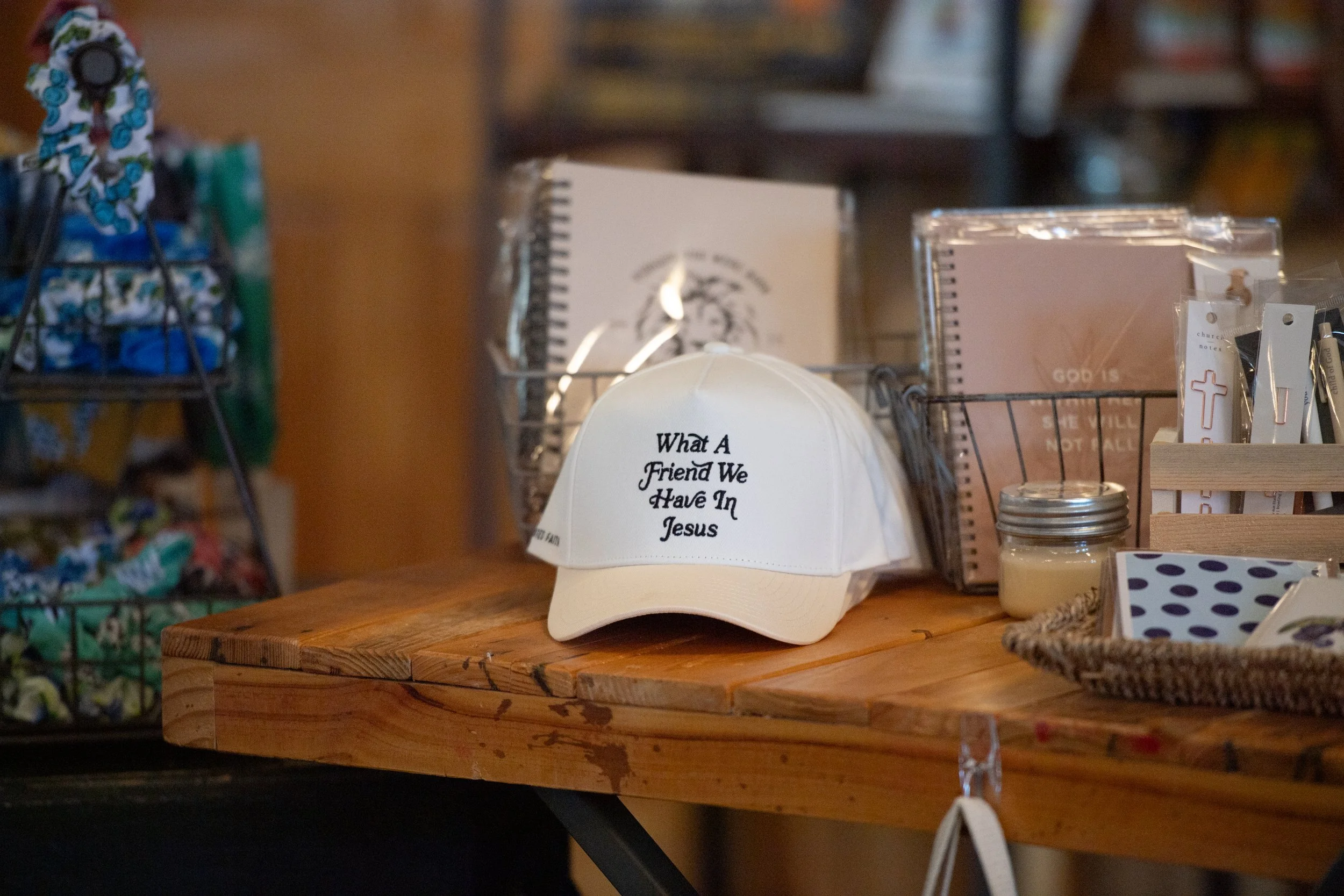 A white baseball cap with the text 'What A Friend We Have In Jesus' printed on it, placed on a wooden table among various religious-themed items.