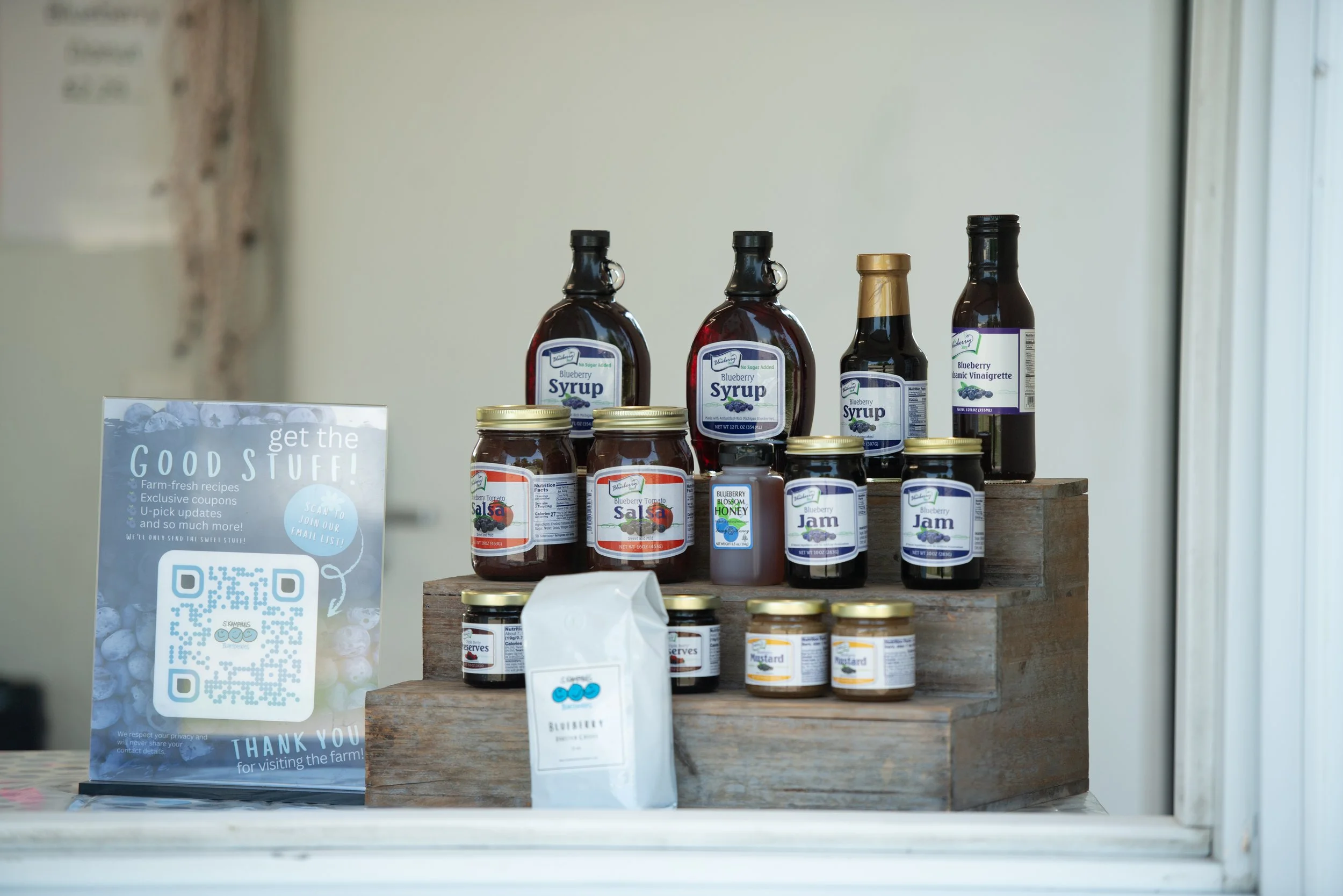 Assorted jars and bottles of blueberry syrup, jam, honey, mustard, and vinaigrette on a wooden display at a farm stand with a sign promoting fresh recipes and coupons.