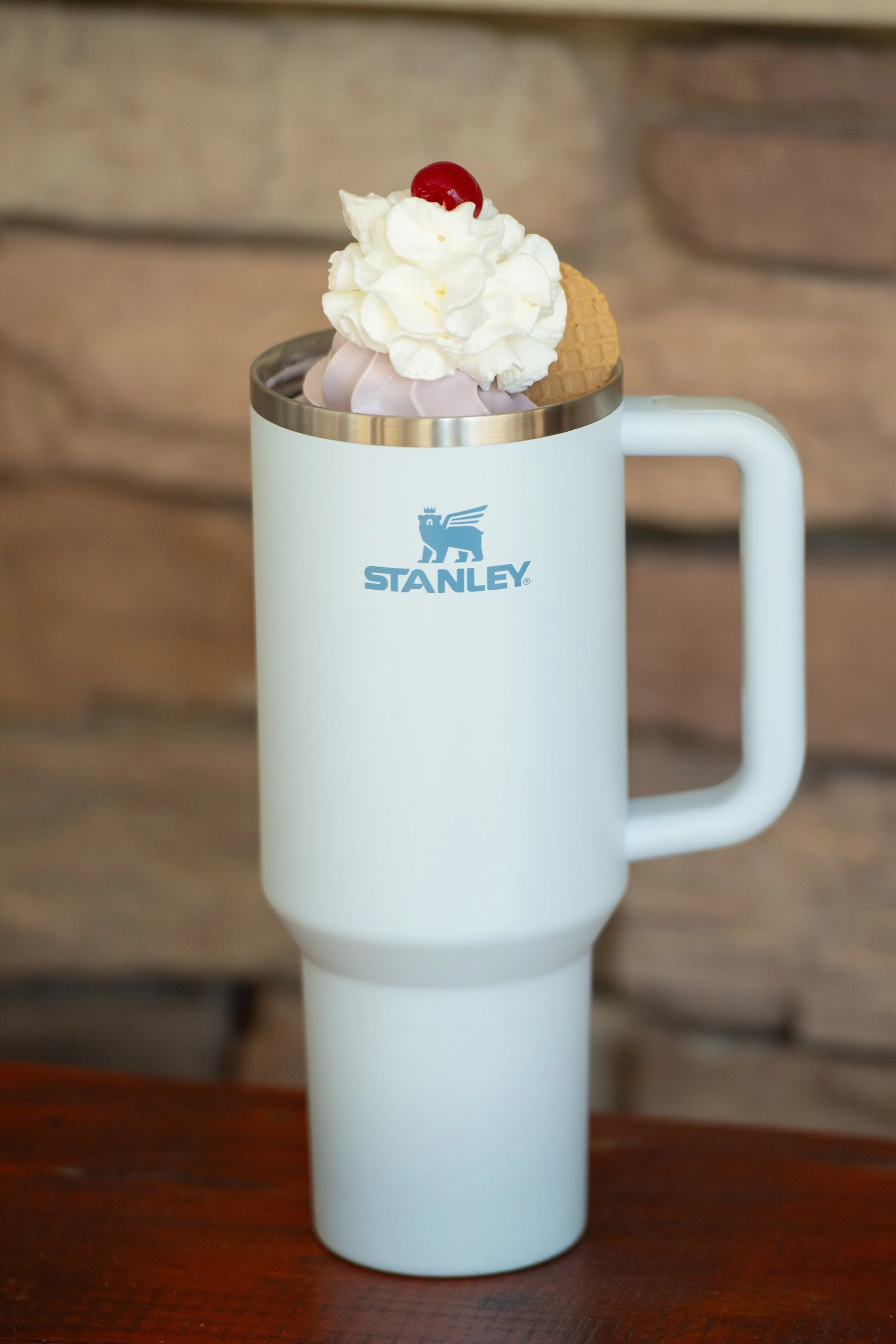 A white Stanley travel mug filled with ice cream topped with whipped cream, a cherry, an ice cream cone, and a splash of pink sauce.