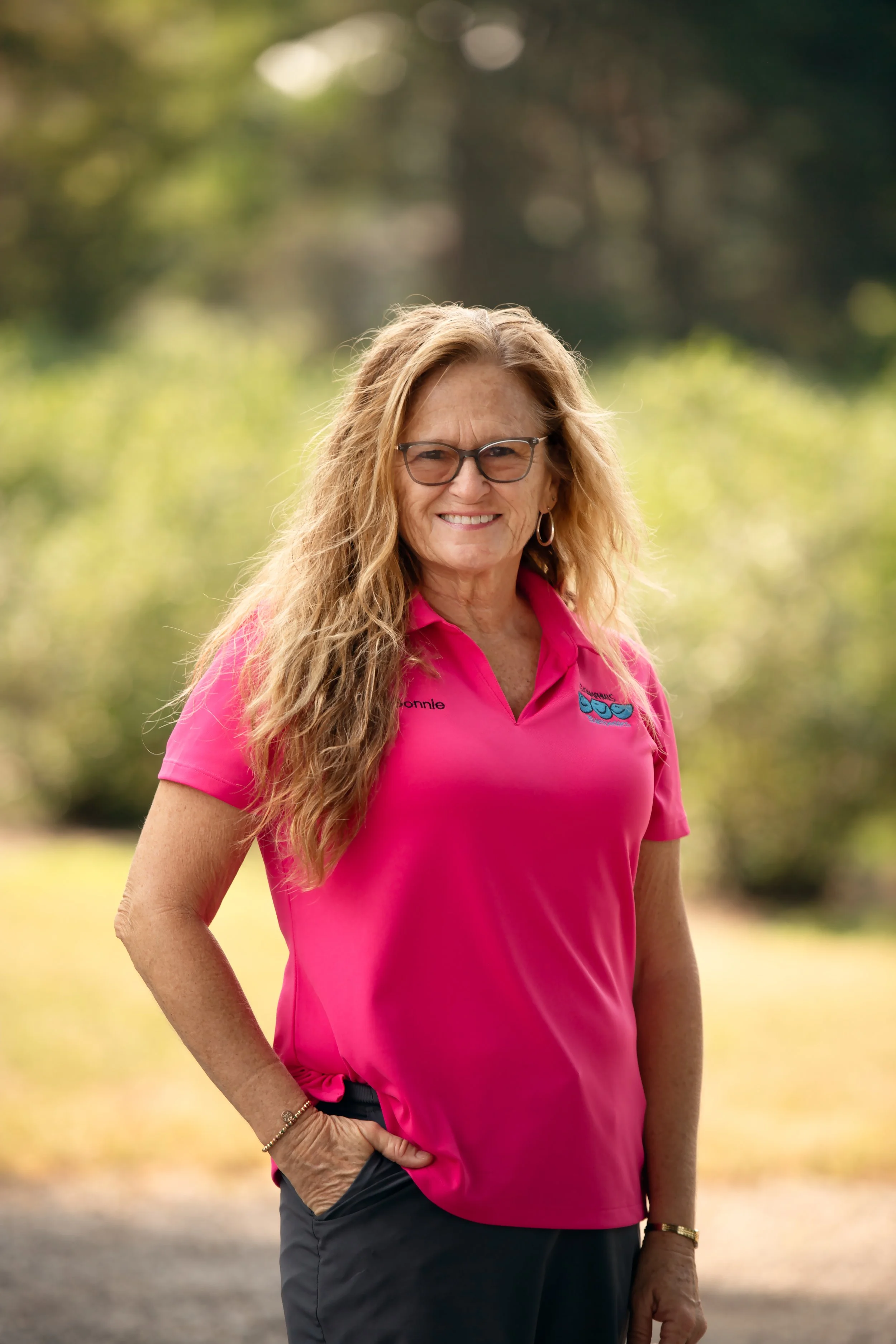 A smiling woman with long, curly, blonde hair, wearing glasses, a pink polo shirt, and dark pants, standing outdoors with a blurred green forest background.