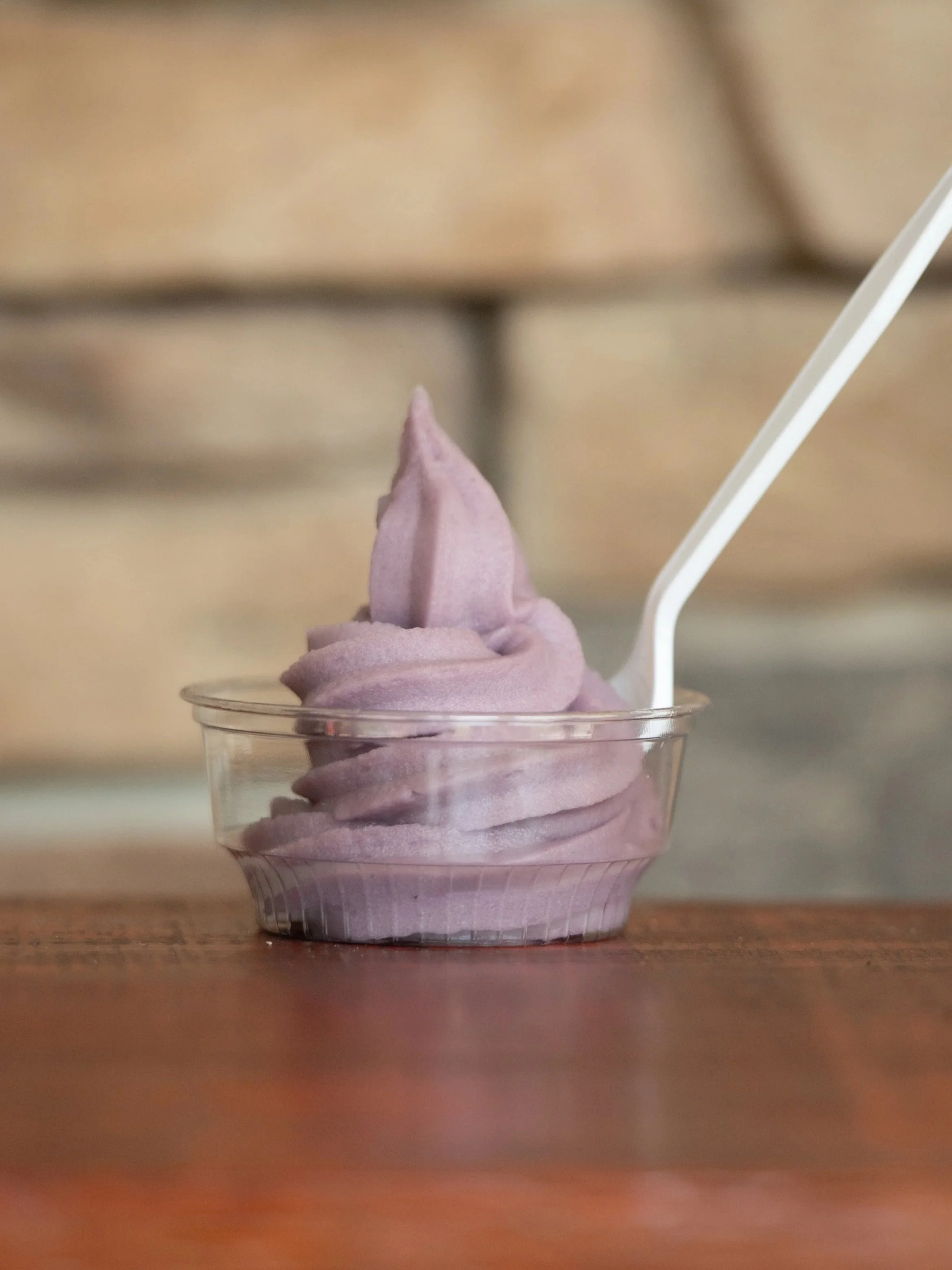 A small clear plastic container of purple soft-serve ice cream with a white spoon on a wooden surface, blurred background.