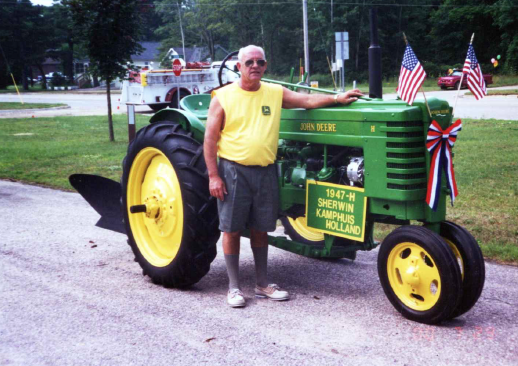 A man standing next to a vintage green and yellow John Deere tractor decorated with American flags and a ribbon, at a parade or event, with a sign indicating it is a 1947 H model from Sherwin Kamphuis in Holland.
