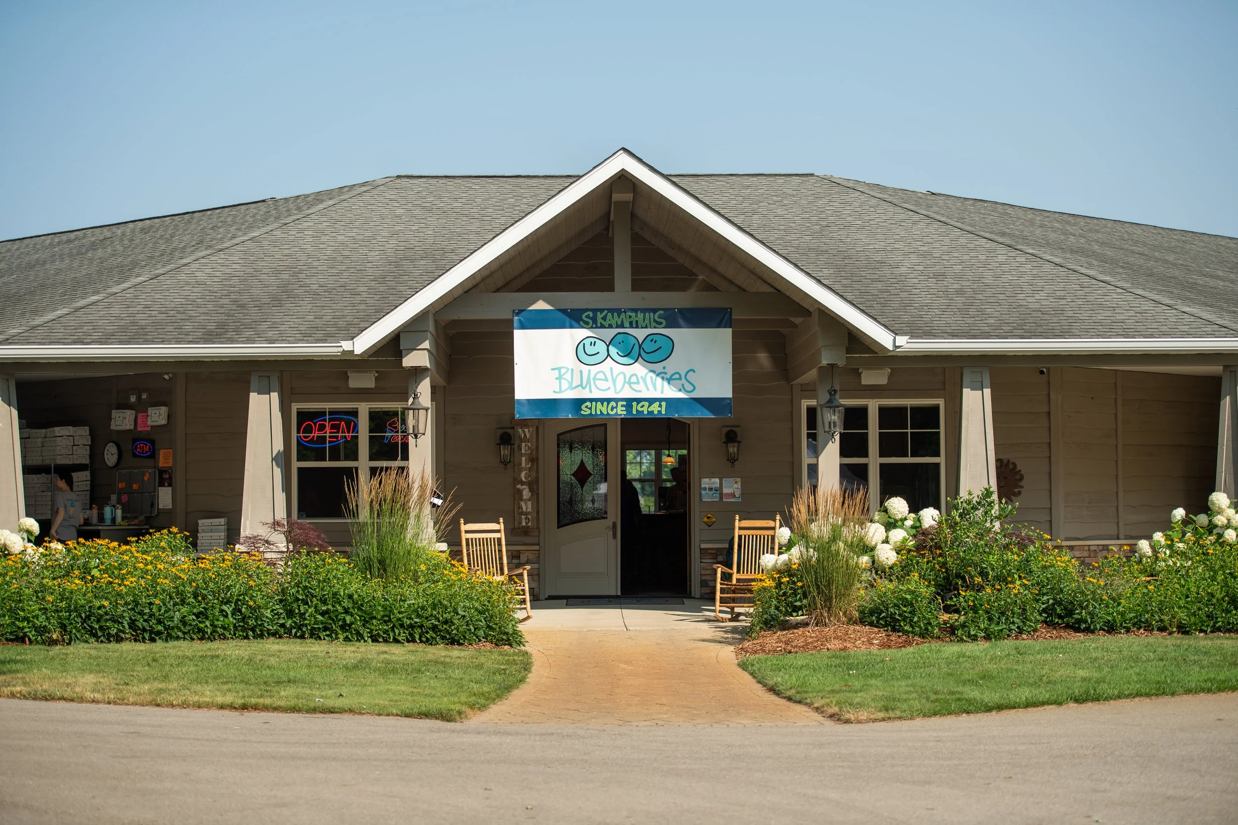 A building with a sign that reads 'Blueberries since 1941' and has a blue smiley face logo. The building has a gray shingled roof, beige siding, and is surrounded by a well-maintained garden with white flowers and green bushes. There is an open sign in the front window and rocking chairs outside the entrance.