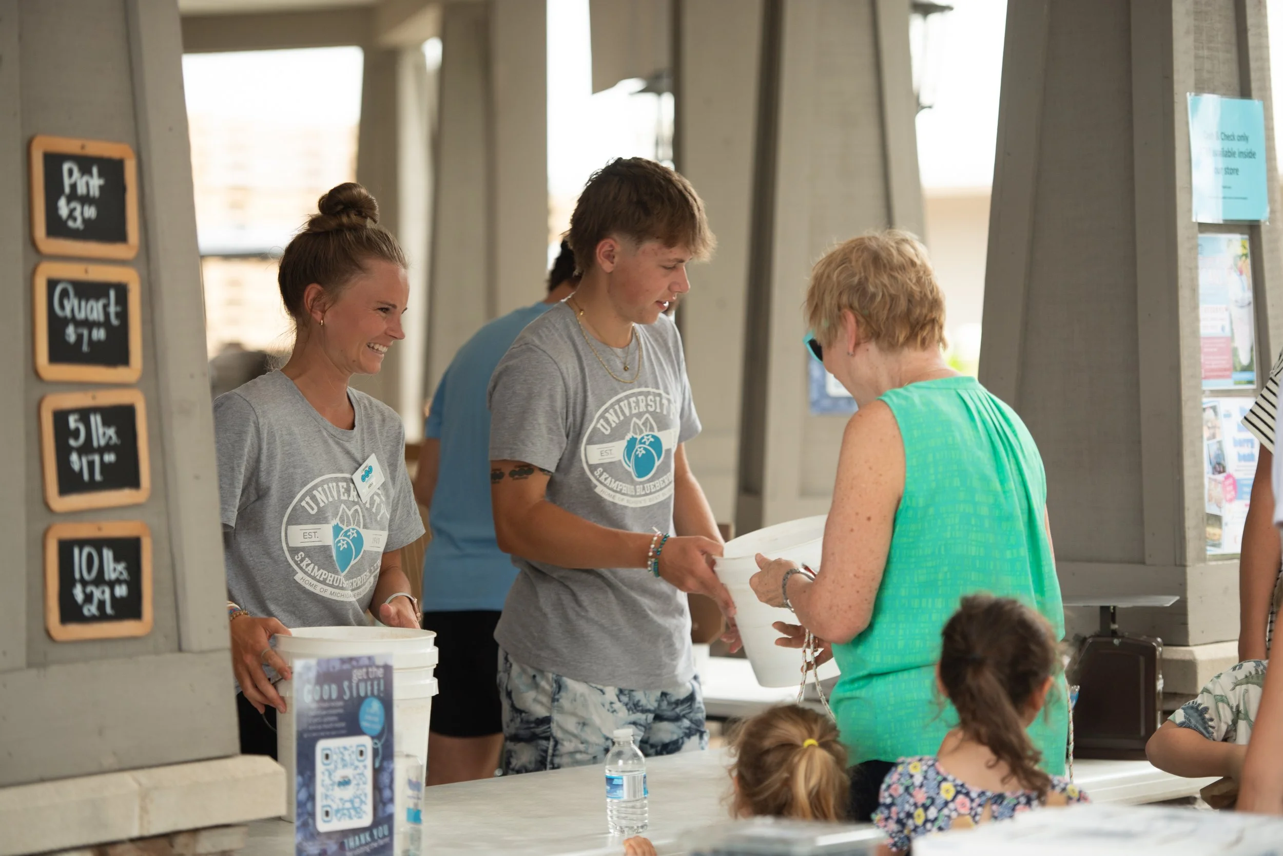 Two young people and an older woman at a food stand, handing over a container to the woman. The young people wear matching gray T-shirts with a logo. There are children in the foreground, and price signs for produce on the side.