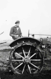 A young boy standing next to an early tractor with large spiked wheels, outdoors in a rural setting.
