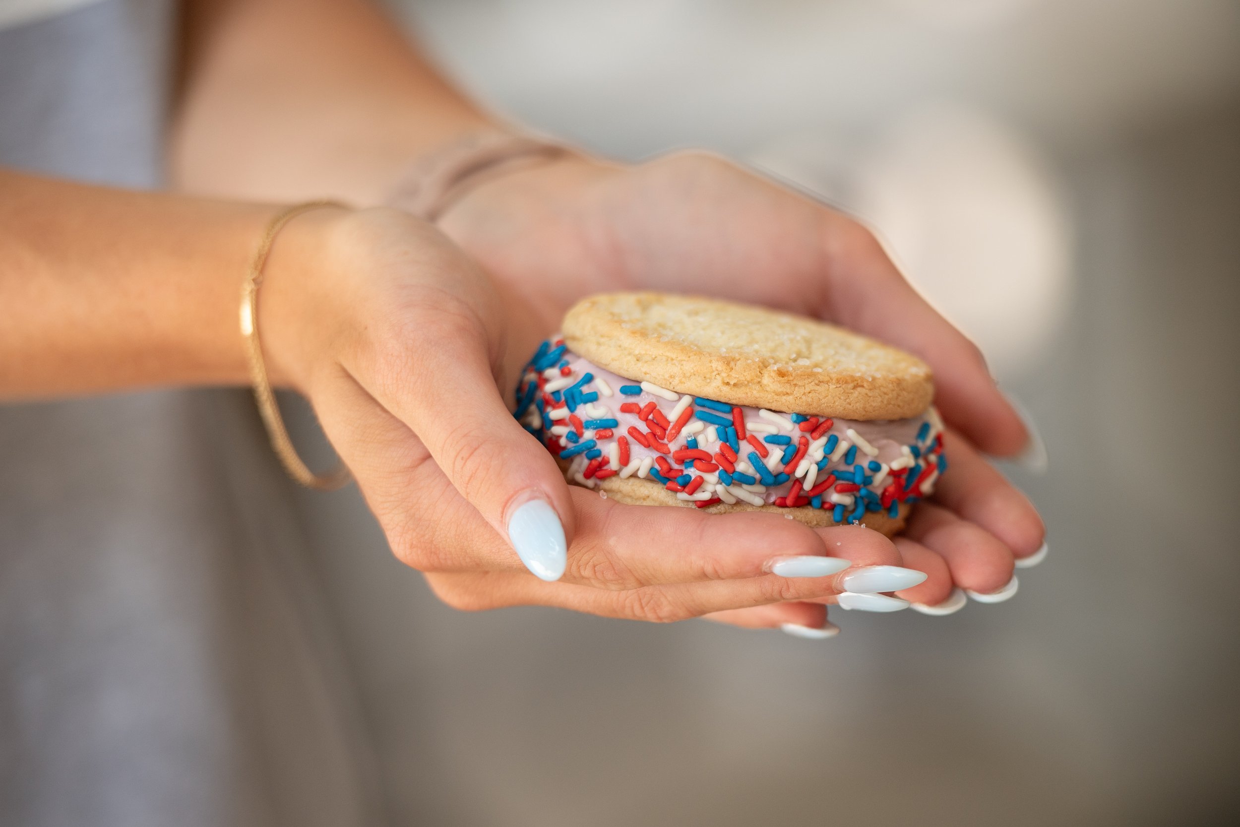 A woman's hands holding a decorated cookie sandwich with red, white, and blue sprinkles and icing, with a cookie on top.