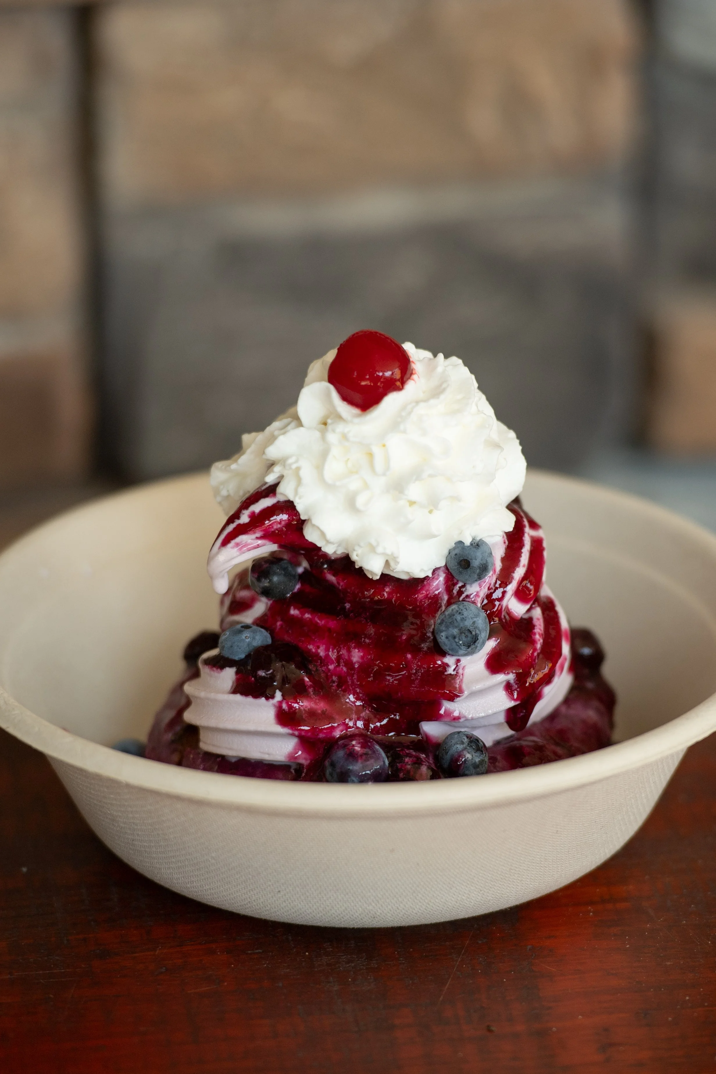 A bowl of blueberry and cherry-flavored soft serve ice cream topped with whipped cream, a cherry, and berry sauce.
