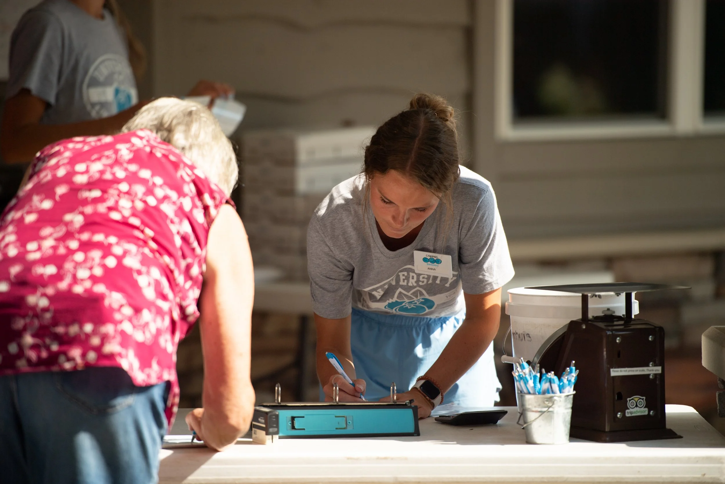 A young woman wearing a gray T-shirt and a smartwatch is signing a document on a clipboard at a registration table, while an elderly woman in a pink floral blouse leans over to sign. There are pens in a container, a scale, and other items on the table.