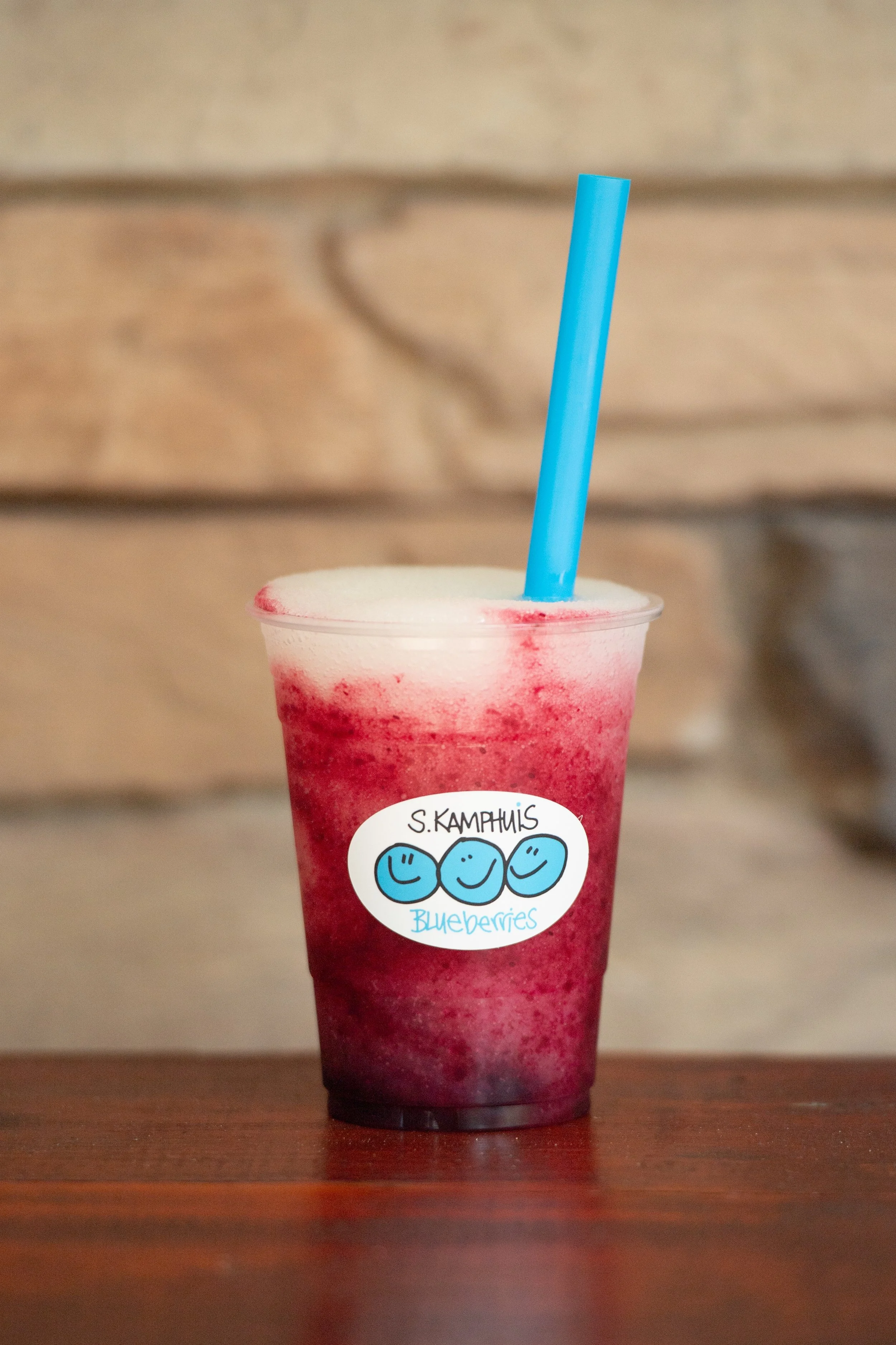 A clear plastic cup filled with a blueberry smoothie with a foam layer on top, a blue straw, and a sticker that says "S. Kamphuis Blueberries" with three smiley faces, placed on a wooden surface against a blurred wooden background.