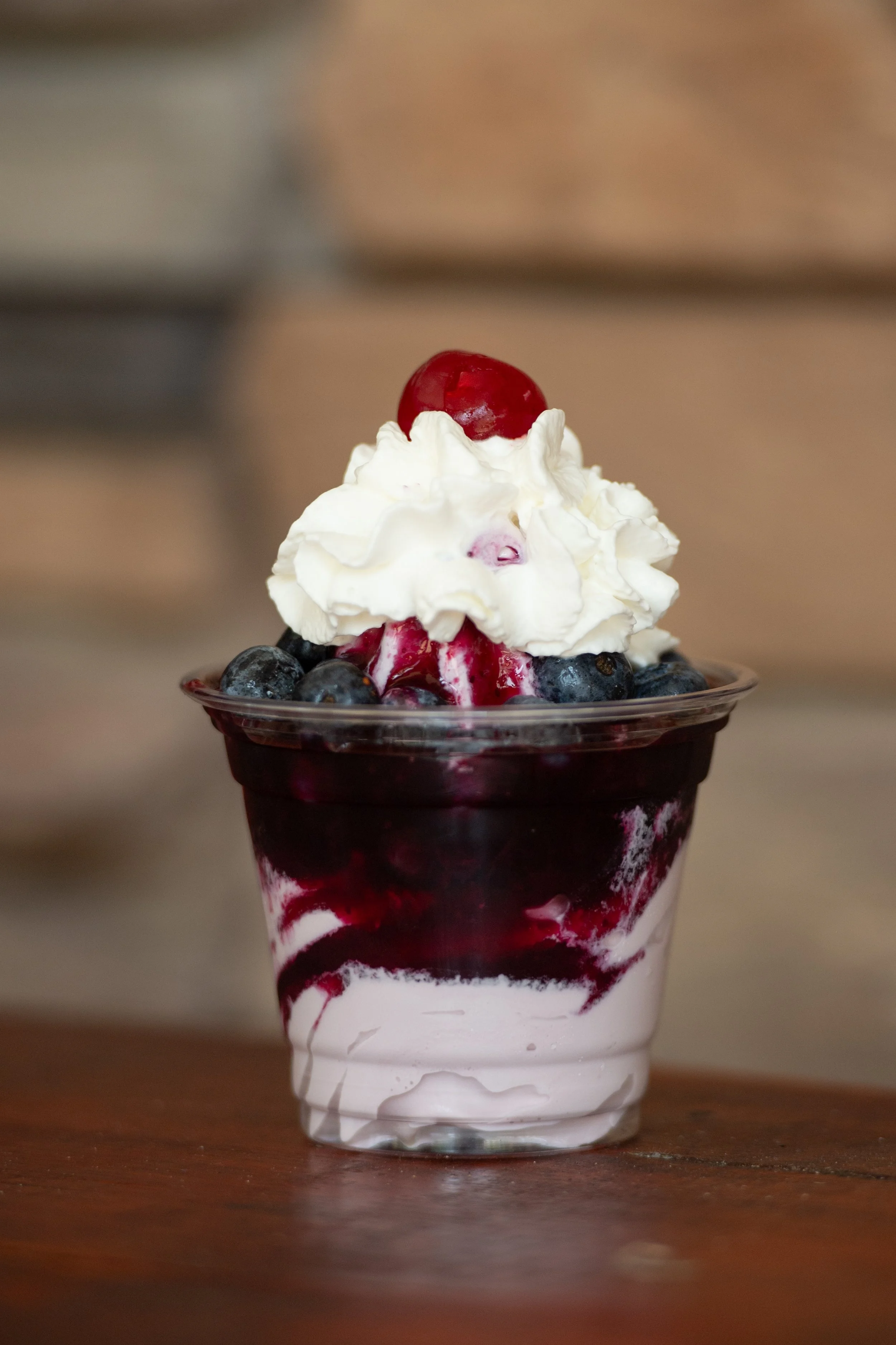 A clear plastic cup filled with layered blueberry dessert topped with whipped cream and a cherry, with blueberries around the whipped cream.