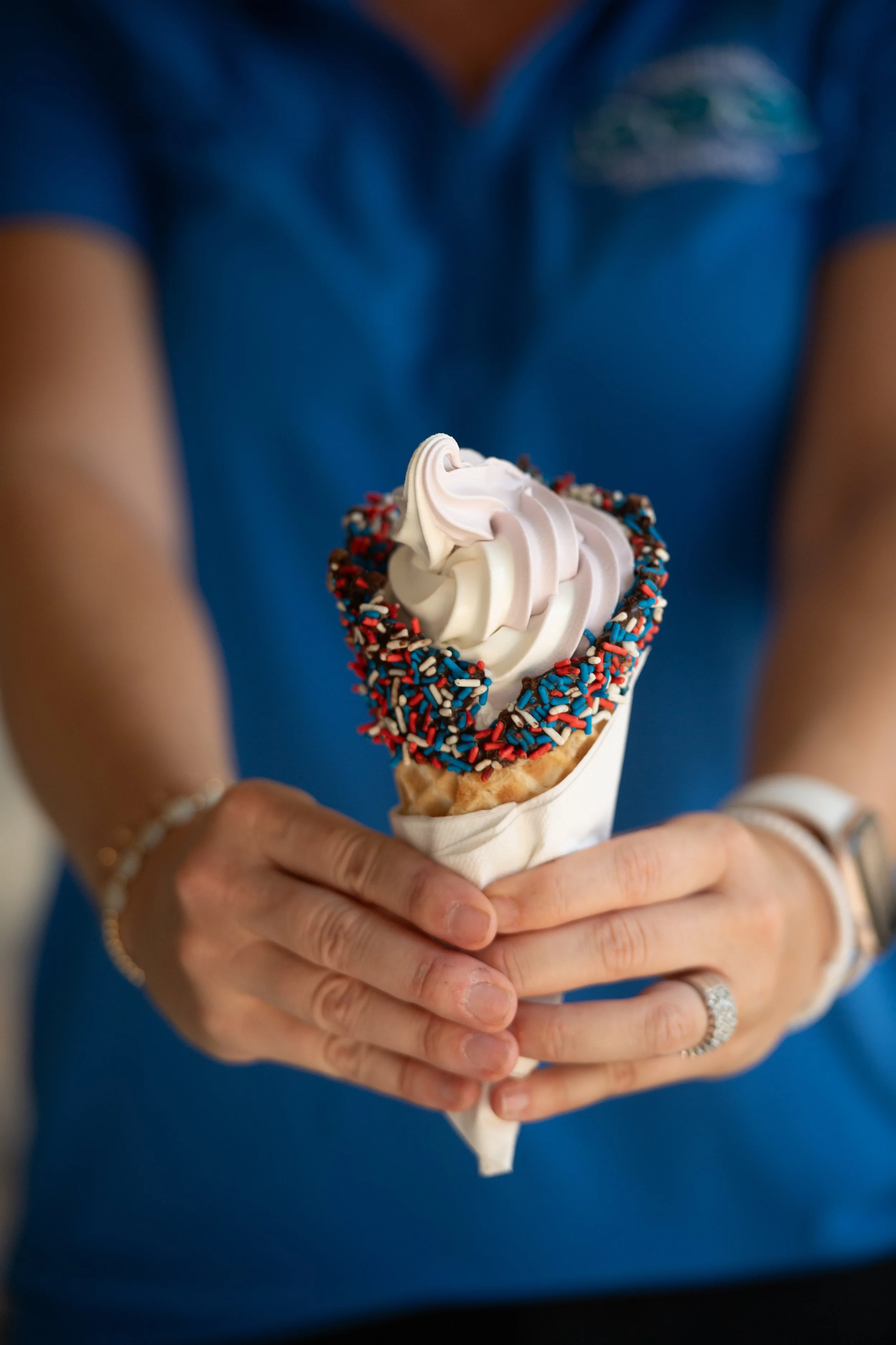 Person holding a soft serve ice cream cone with a red, white, and blue sprinkles border, in front of a blurred person wearing a blue shirt.