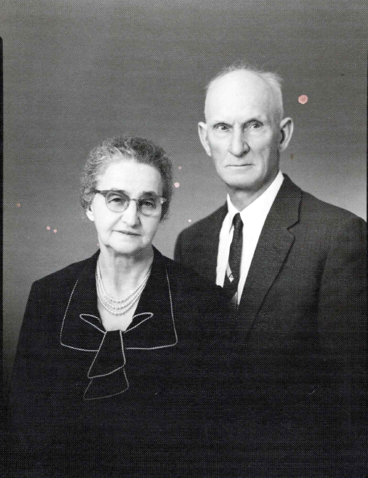A black and white photo of an elderly woman and man standing together, both dressed formally. The woman is wearing glasses, a dark blazer with a ribbon detail, and a pearl necklace. The man is wearing a dark suit and tie, with a serious expression.