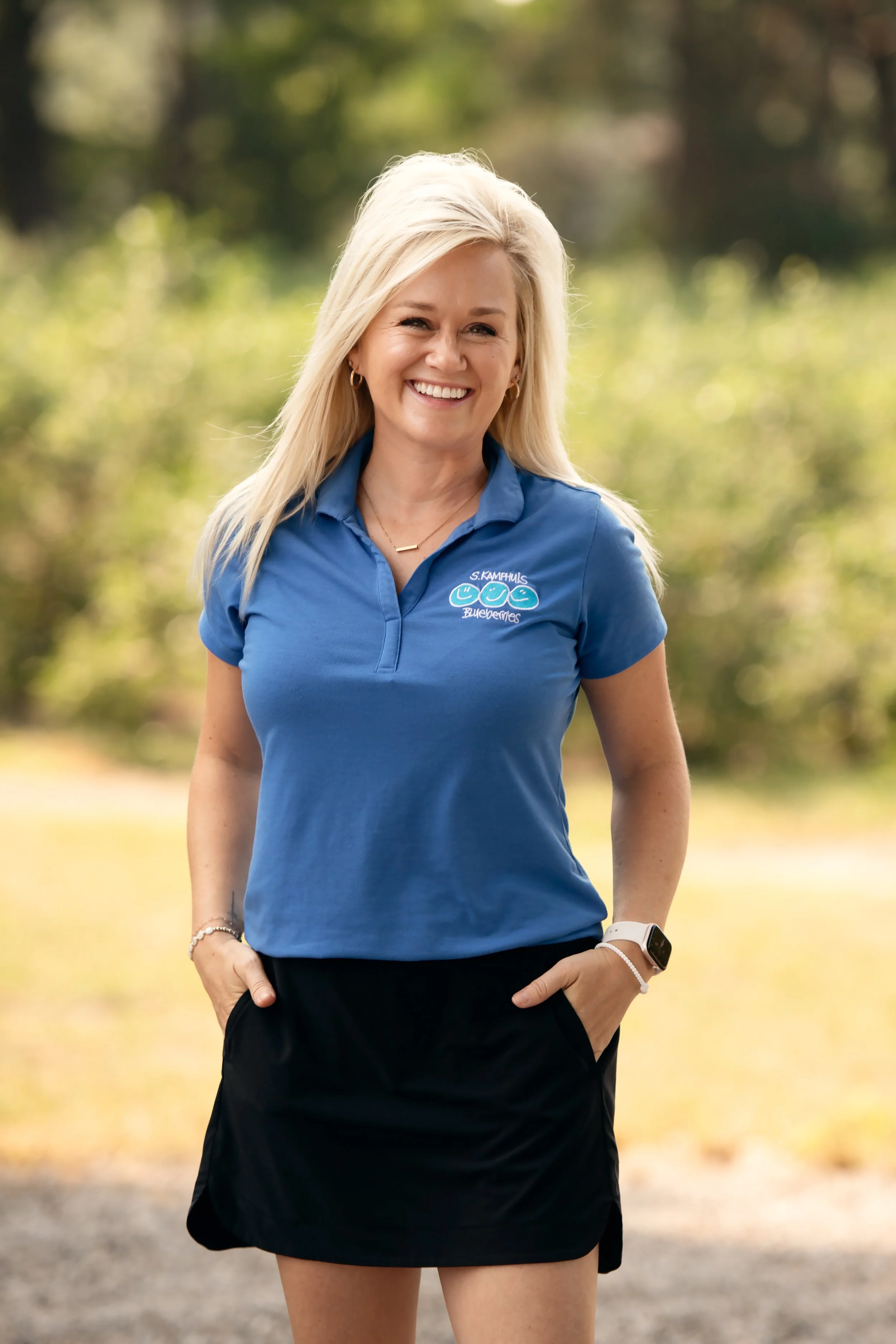 A smiling woman in a blue polo shirt and black skirt standing outdoors with hands in pockets, surrounded by a blurred natural background.