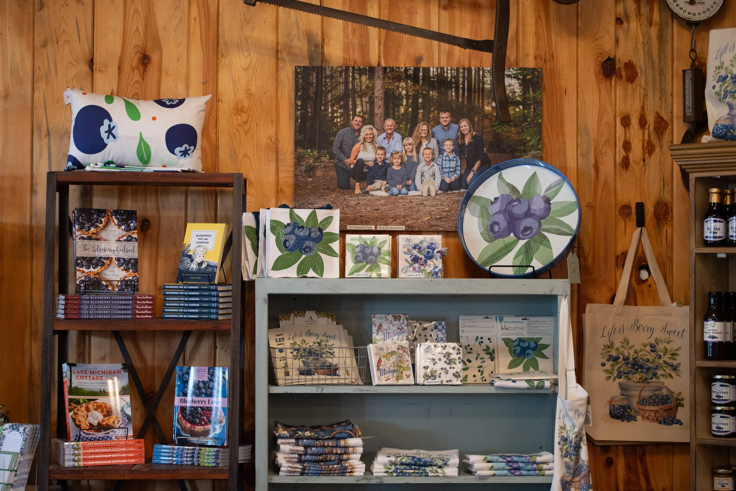 Display of blueberry-themed books, prints, and textiles on wooden shelves with a family portrait hanging on a wood-paneled wall.