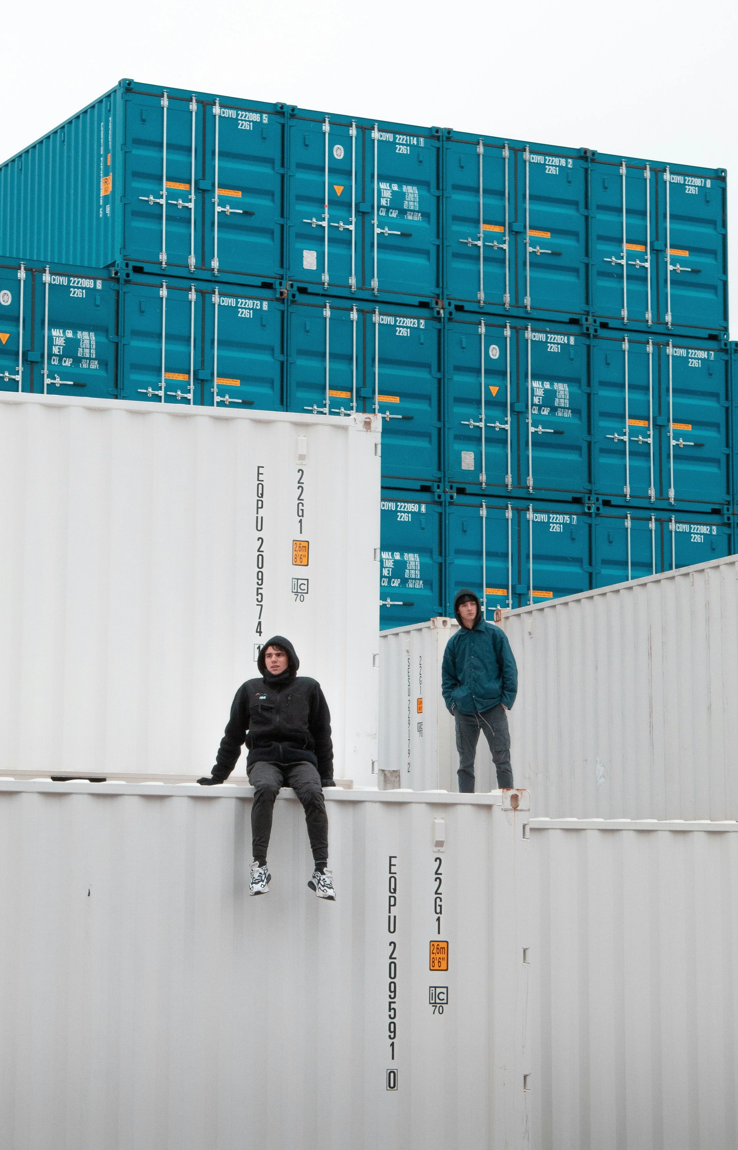 Two people sitting and standing on stacked cargo containers, with blue and white containers around them, in an industrial setting.