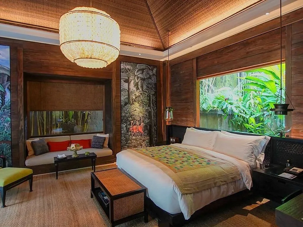 A bedroom with a large bed, bedside tables, and tropical-themed decor. There is a sofa by the window, large windows with lush greenery outside, and warm wooden walls and ceiling with a large chandelier overhead.