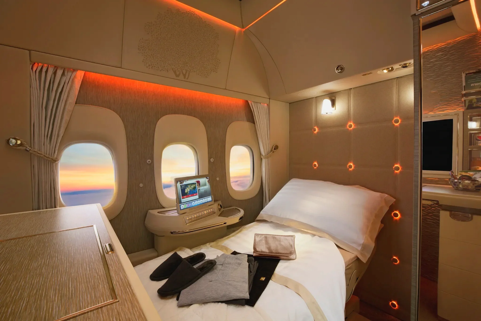 Luxurious first class airplane suite with a bed, a sunset view, and warm yellow and orange lighting accents.