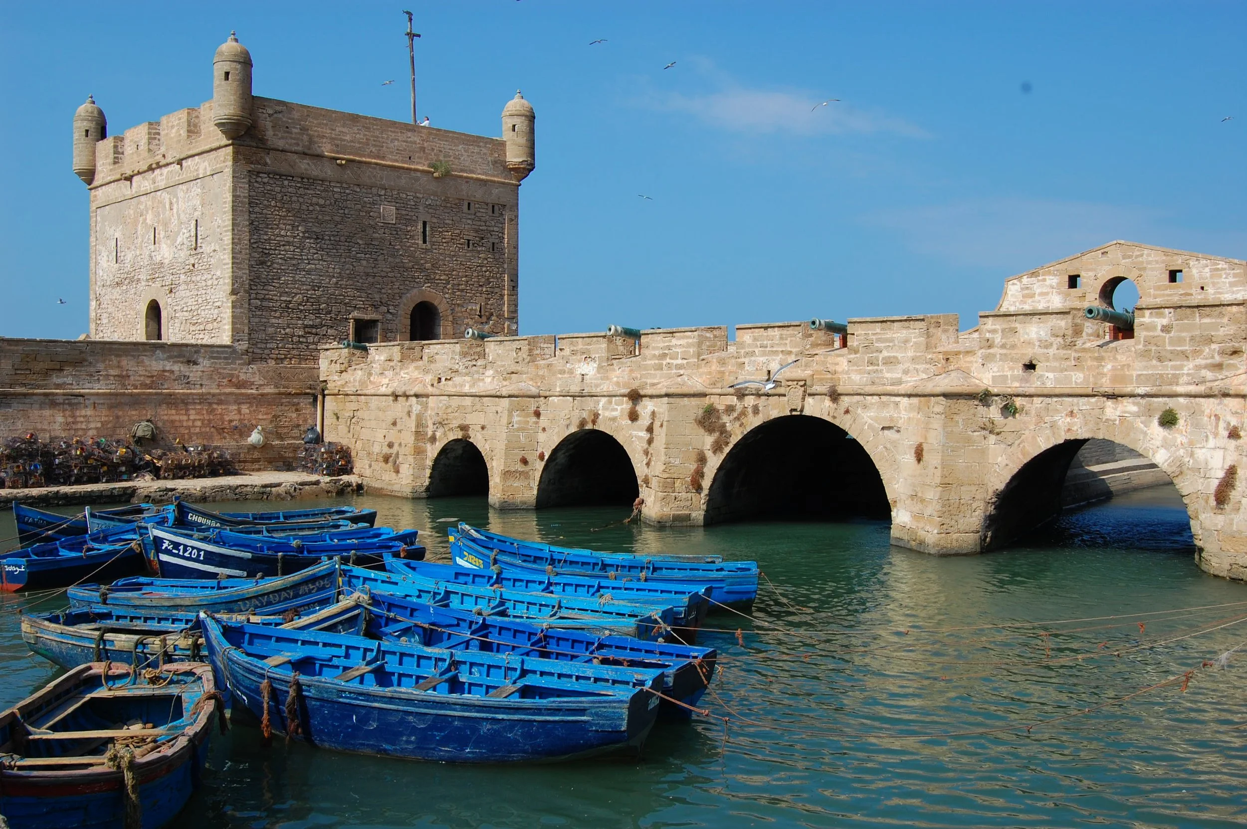 A historic stone fortress with a tower, arched bridge, and cannons, next to a body of water with blue boats anchored nearby, under a clear blue sky.