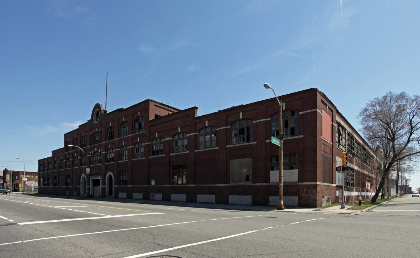 A large, abandoned red-brick building on a corner with broken windows, an empty parking lot in front, and a tree on the sidewalk. The sky is clear and blue.