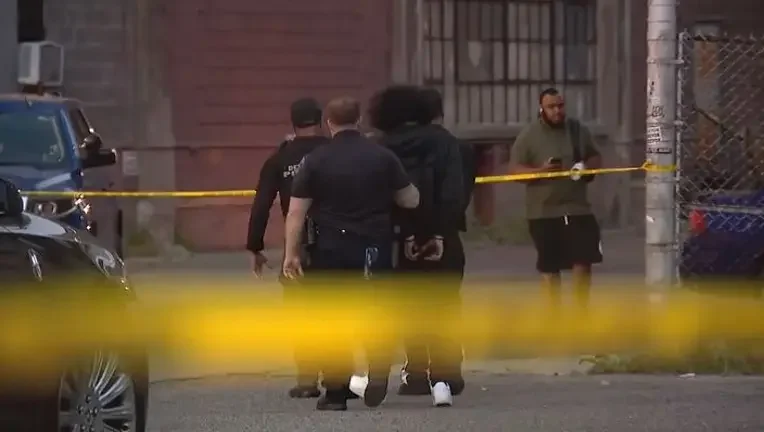 Police officers and civilians at a crime scene behind yellow caution tape, with police and civilian vehicles nearby.
