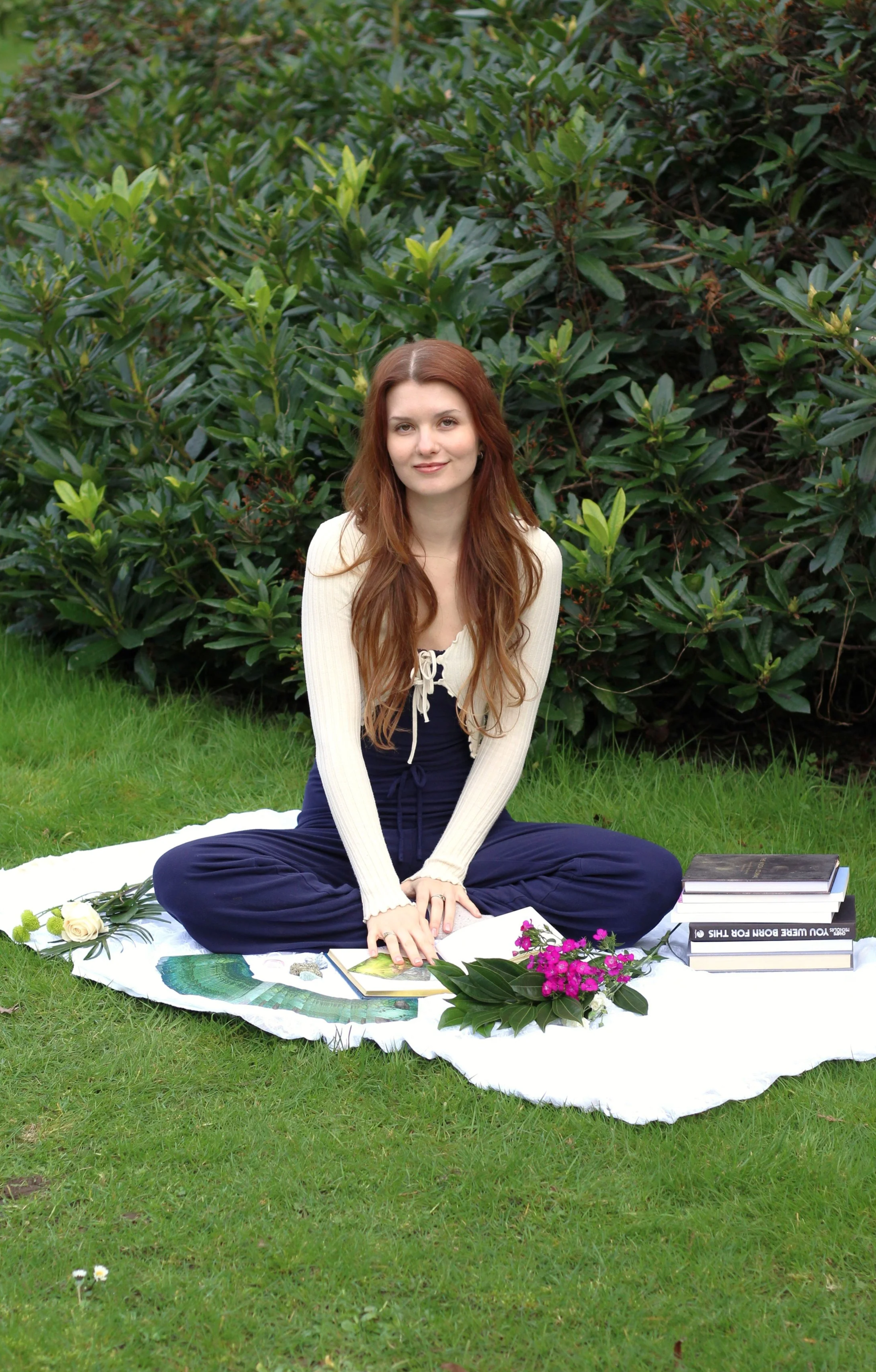 A young woman with long red hair sits cross-legged on a white sheet on the grass in front of green leafy bushes. She is smiling softly and wearing a cream-colored long-sleeve top and dark blue pants. There are books, flowers, and artwork around her, suggesting she is engaged in reading or artistic activities outdoors.