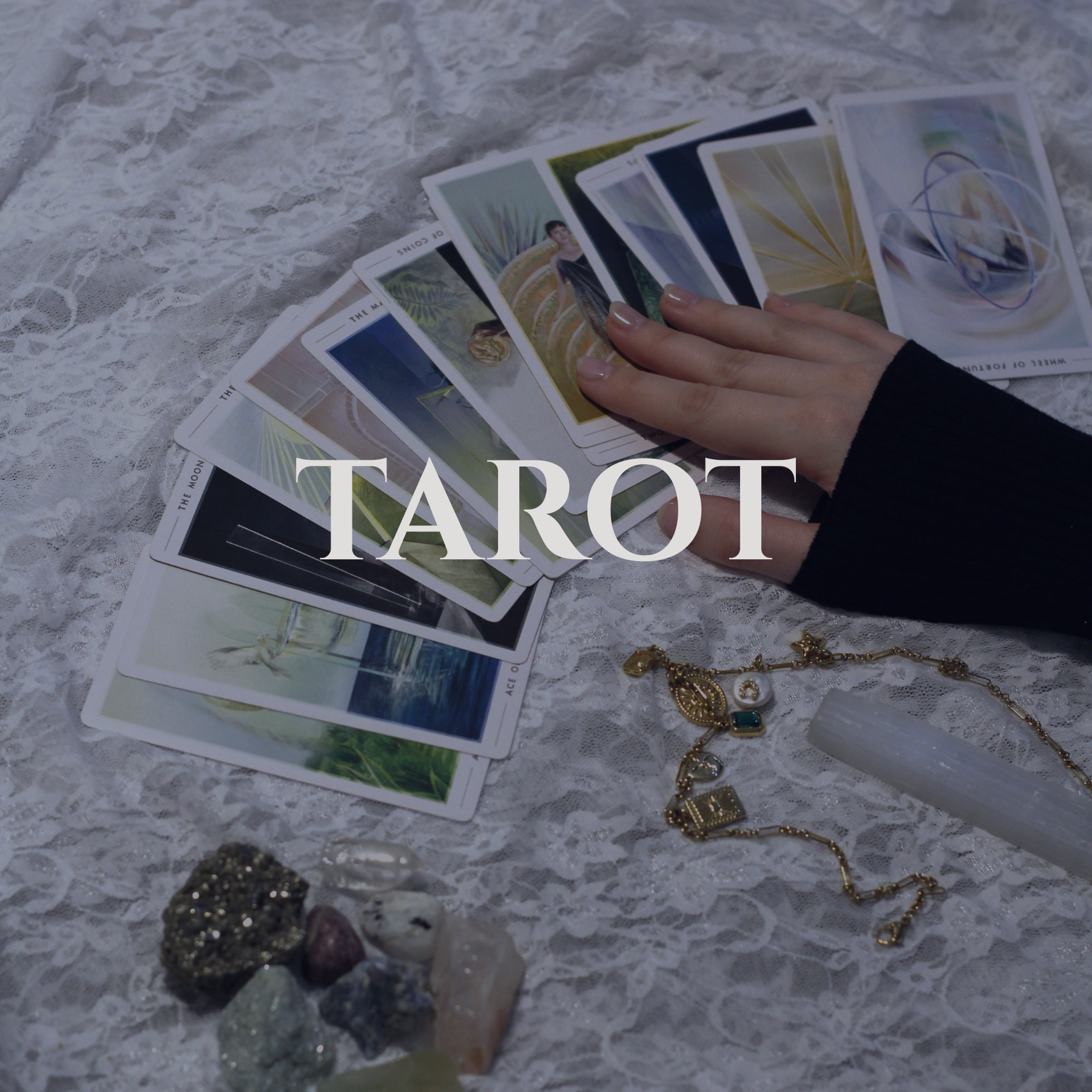 A hand with light-colored nail polish holding tarot cards spread out on a lace surface, with jewelry and a ring nearby.