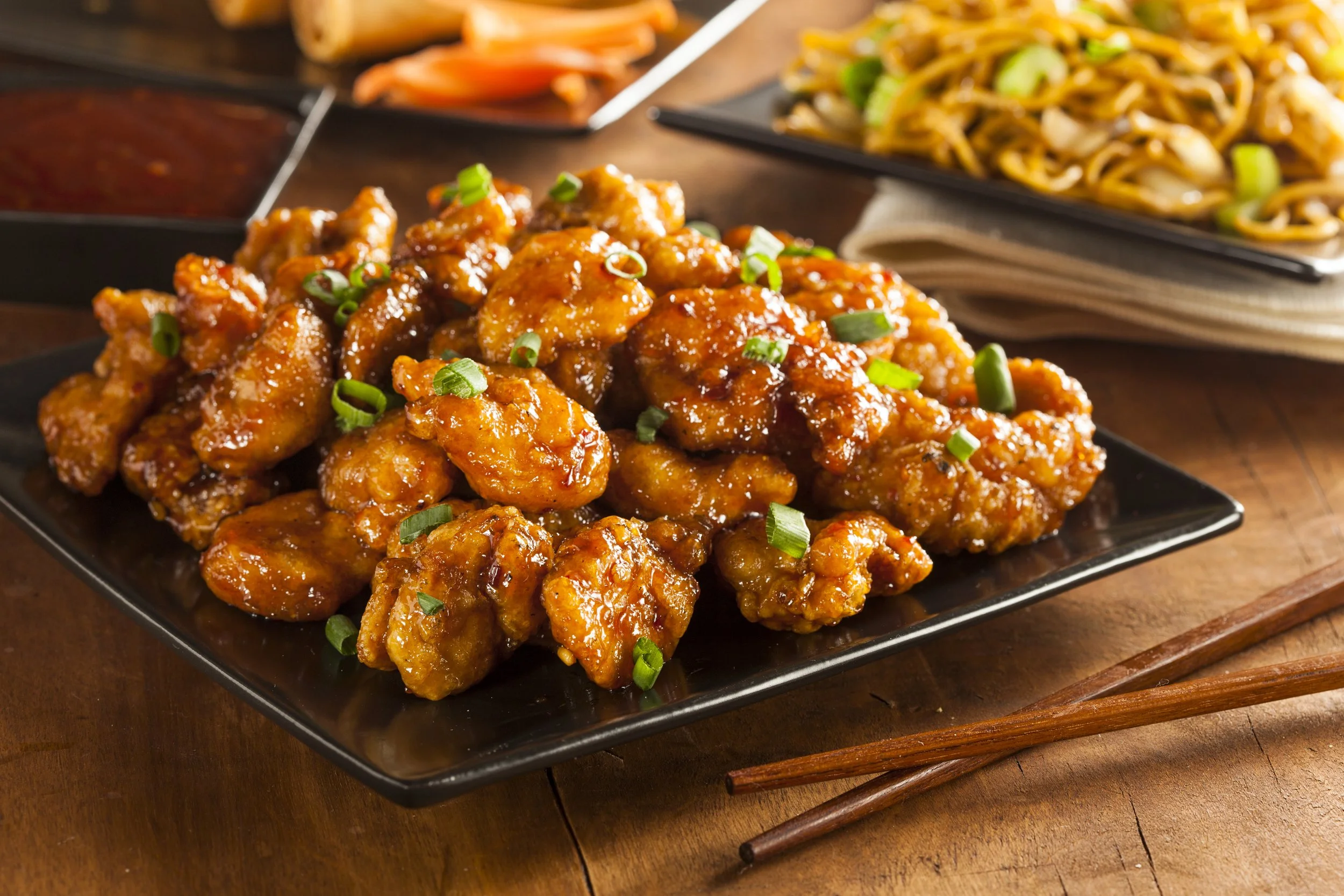 Plate of crispy orange chicken garnished with chopped green onions, with side dishes of stir-fried noodles and sauces in the background.