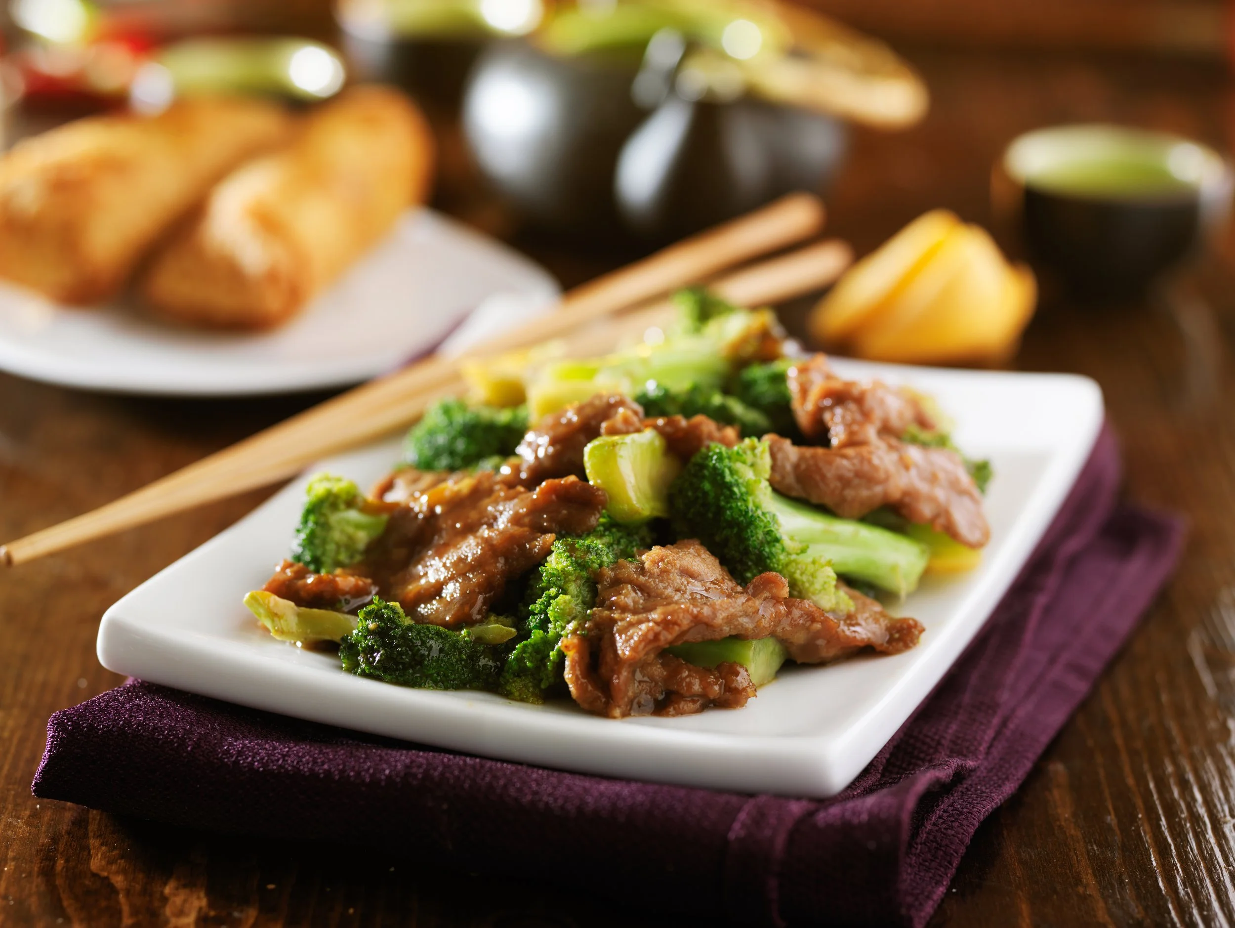 Stir-fried broccoli and beef on a white plate, placed on a purple napkin, with a plate of spring rolls and dipping sauces in the background.