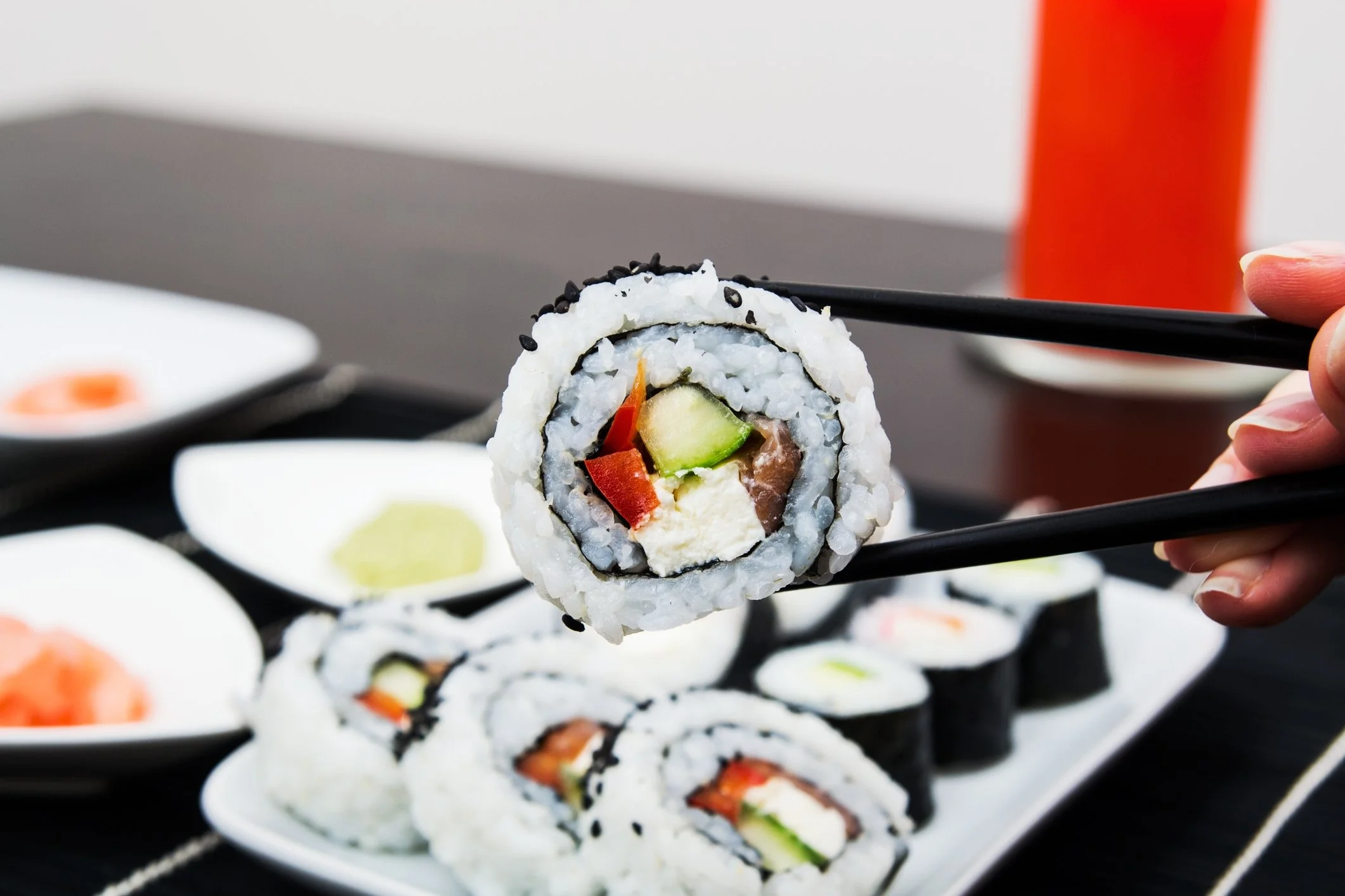 Hand holding chopsticks picking up a sushi roll with cucumber, red pepper, cream cheese, and fish, on a plate of assorted sushi in the background.
