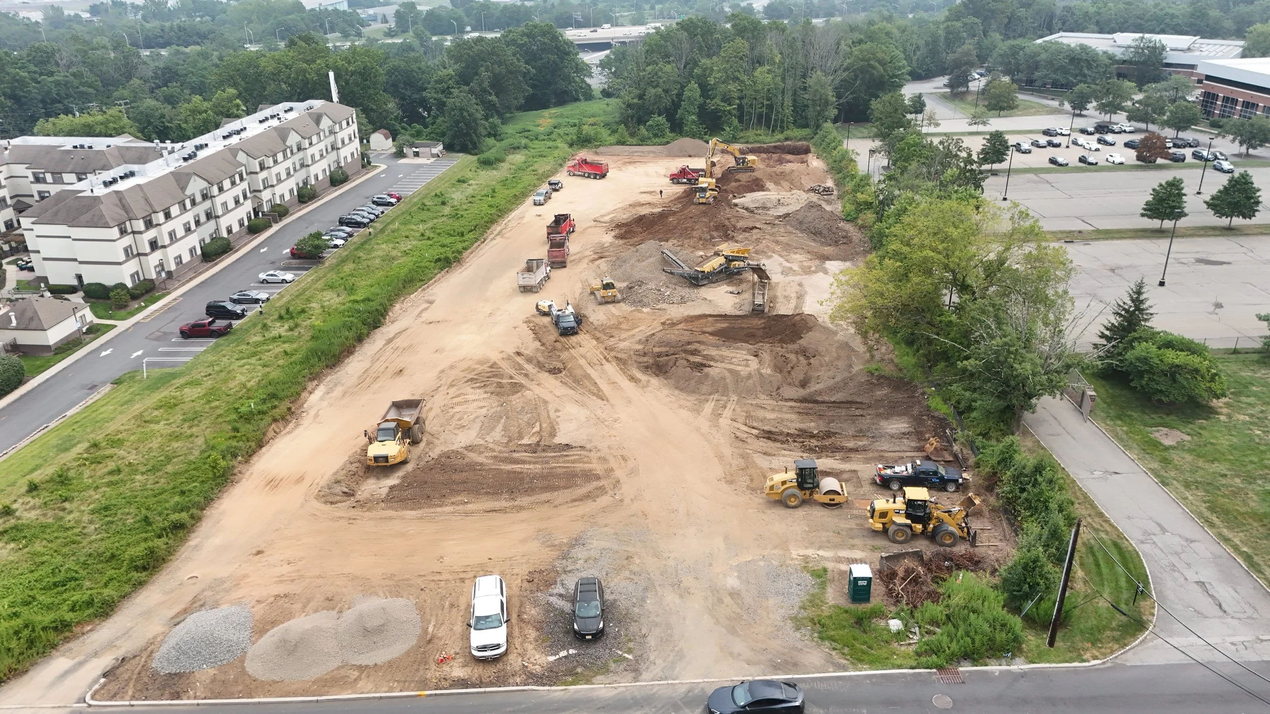 Construction site with various machinery and vehicles, adjacent to a residential area and parking lot, in a green landscape.