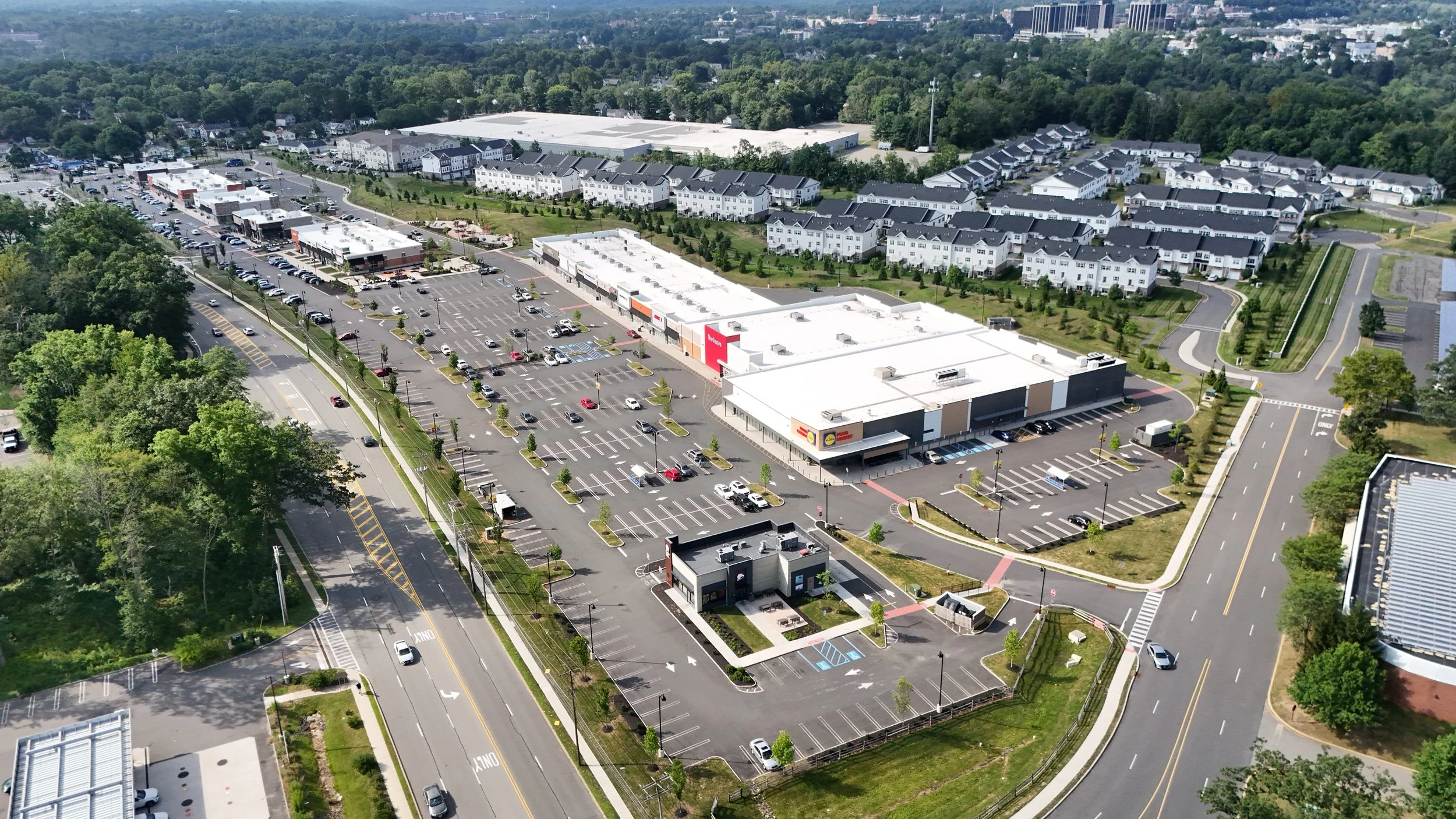 Aerial view of a shopping plaza with parking lots, retail stores, and surrounding residential area with trees and houses.