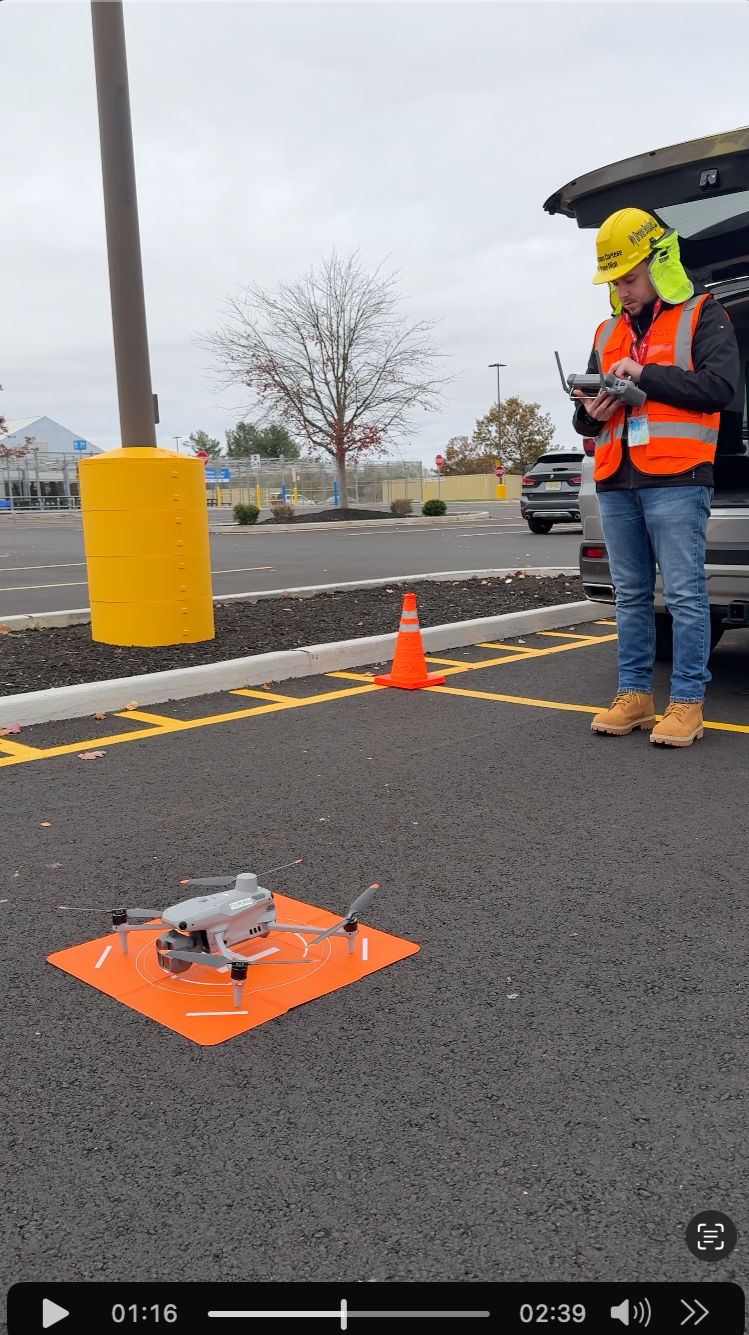 A man in safety gear, including a yellow helmet and orange vest, is standing in a parking lot next to an open car trunk and a drone on an orange landing pad. The man appears to be operating or inspecting the drone.