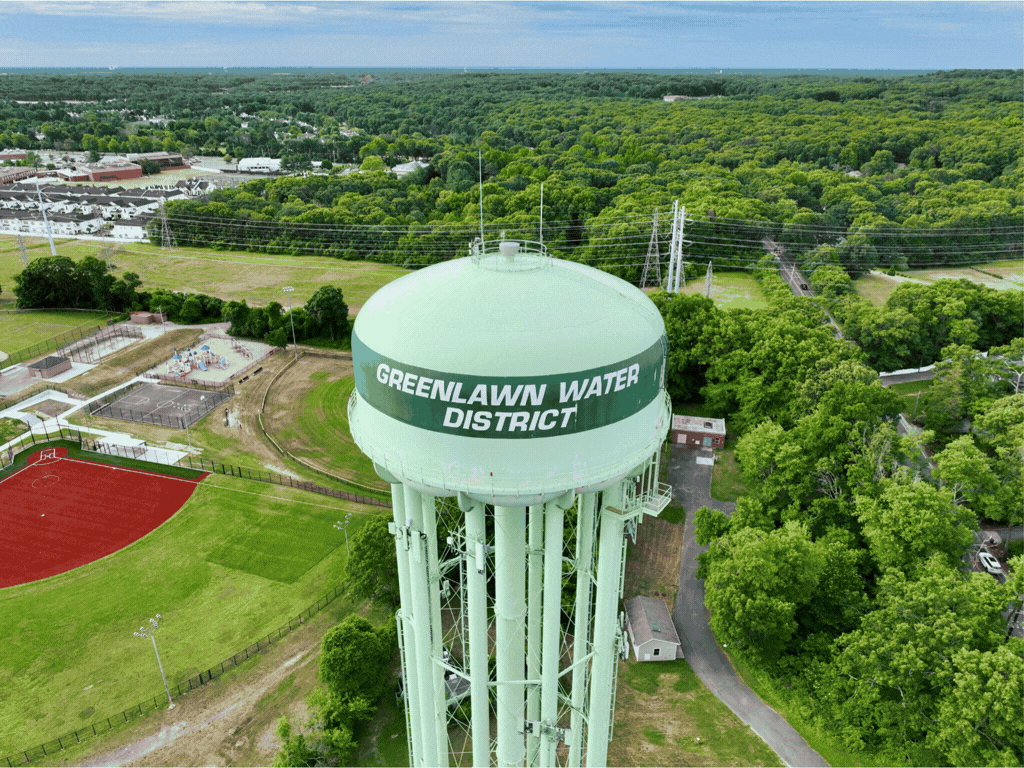A large water tower with green paint and the words 'Greenlawn Water District' written on it, situated above a park with sports fields and a wooded area in the background.