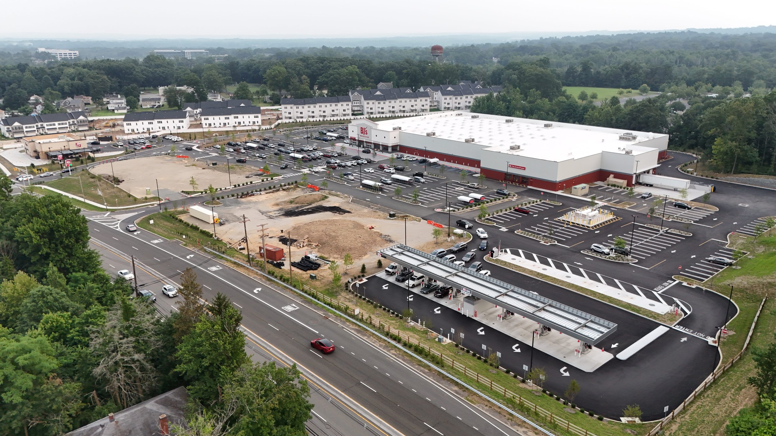 An aerial view of a shopping center with a large parking lot, some construction work ongoing in one area, and a gas station with multiple pumps in the foreground. Surrounding greenery and residential buildings are visible in the background.