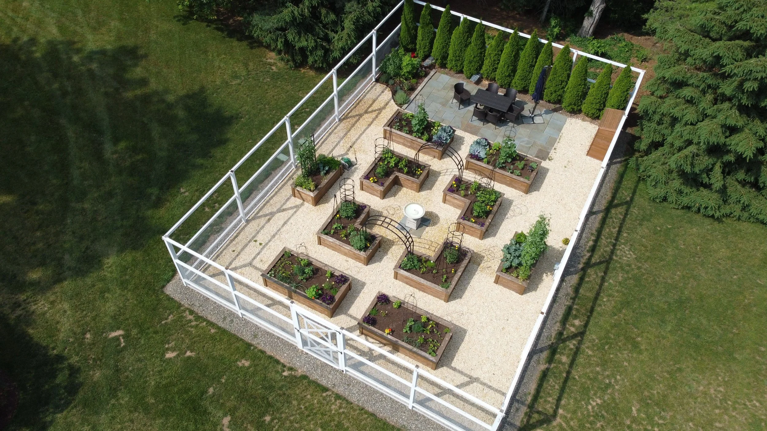 A fenced backyard garden with raised wooden garden beds filled with plants, a gravel pathway, a white birdbath in the center, and a patio area with a table and chairs surrounded by lush trees.