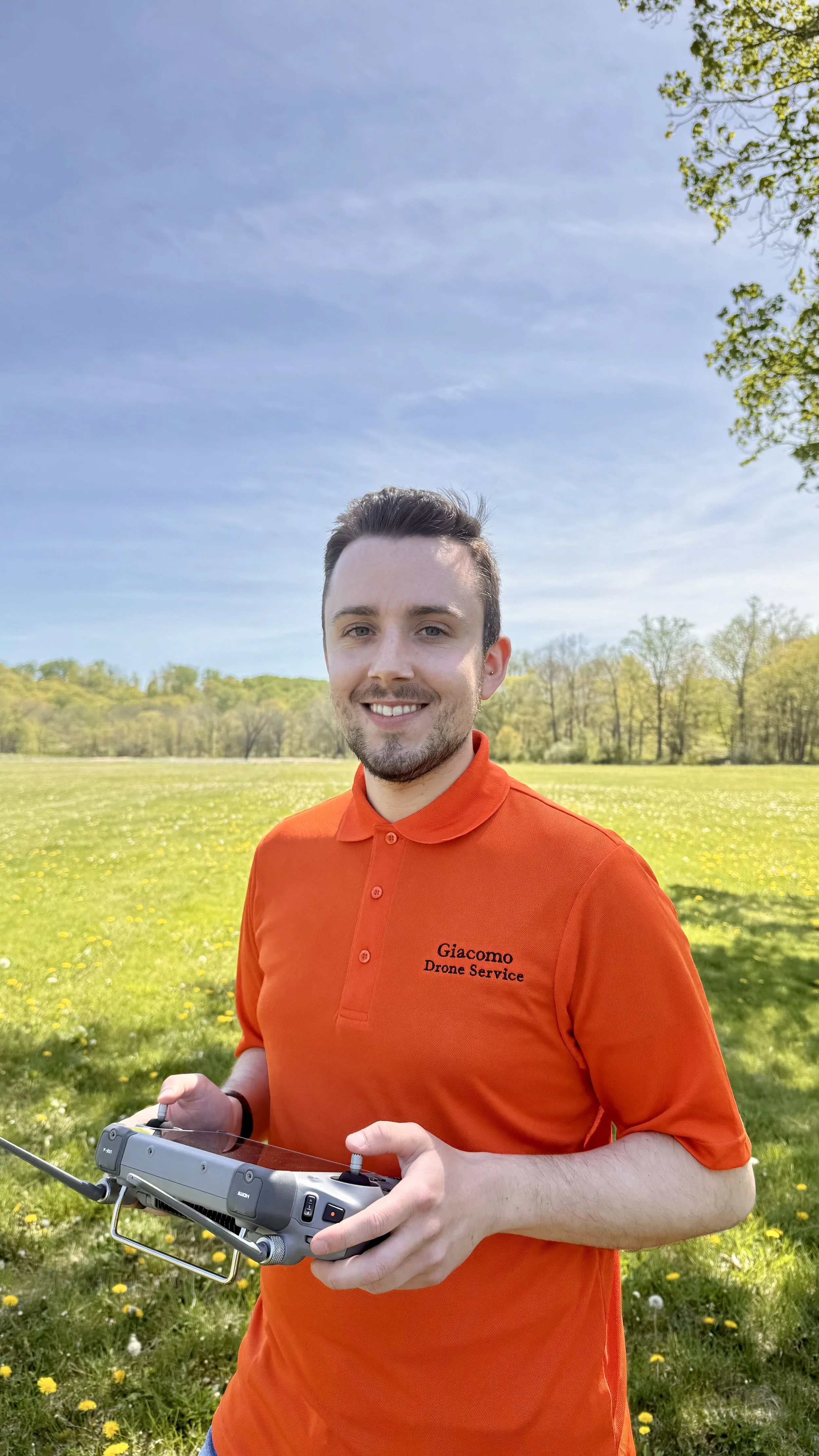A young man with short dark hair, a beard, and light skin, smiling while holding a drone controller outdoors in a grassy park with trees and a blue sky in the background. He is wearing a bright orange polo shirt with a logo that reads 'Giacomo Drone Service'.