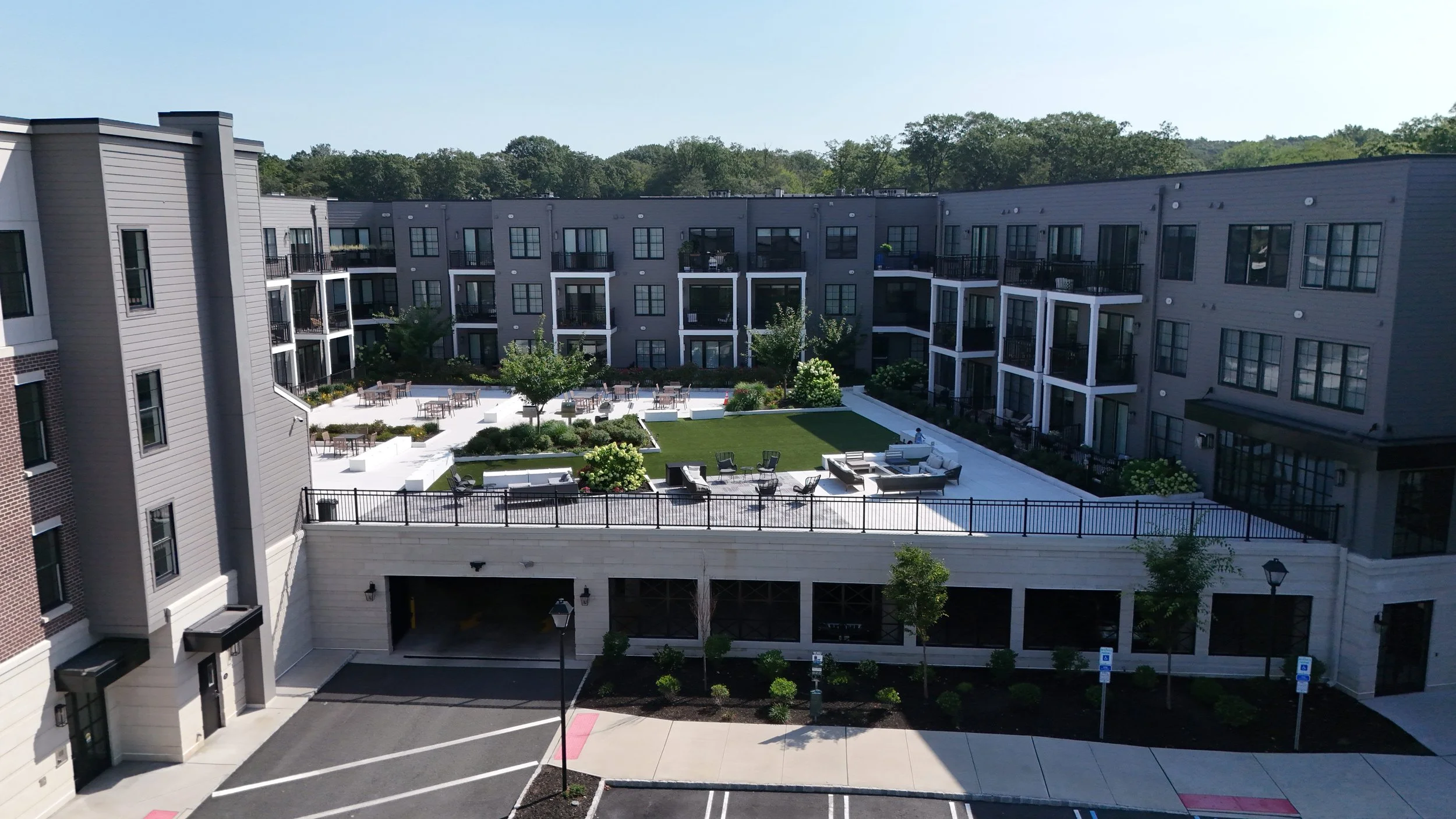 A modern apartment complex with a rooftop patio, outdoor seating, and landscaped gardens, viewed from an elevated perspective.