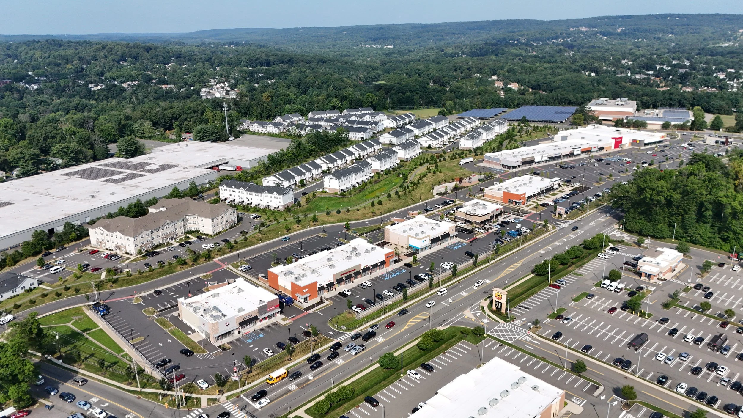 Aerial view of a commercial shopping plaza with parking lots, retail stores, and a large retail building, surrounded by residential neighborhoods and green hills in the background.