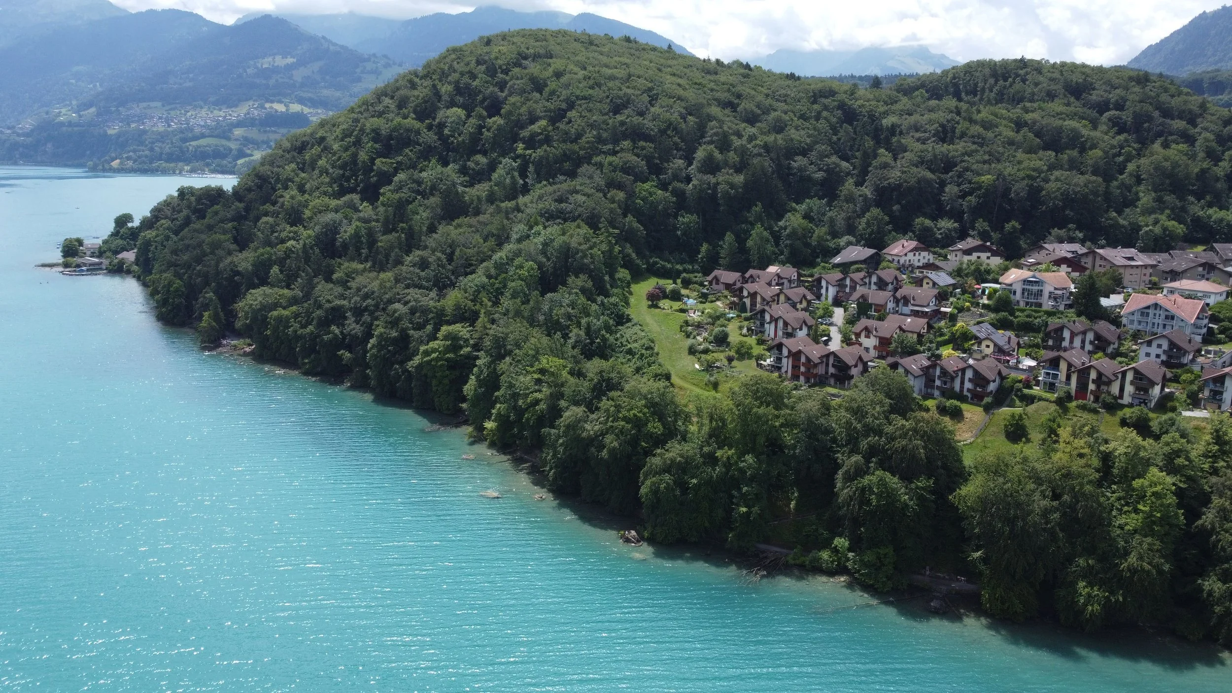 Aerial view of a lakeside residential area with houses on a green hillside beside a calm, turquoise lake with mountainous background.